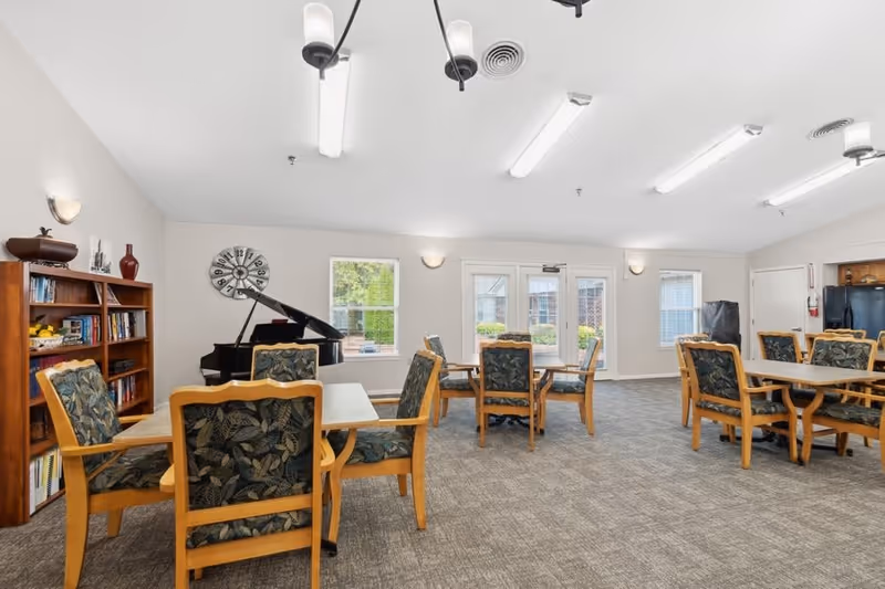 A spacious common room with multiple tables and chairs featuring floral upholstery. There is a black grand piano near the back wall, a wooden bookshelf filled with books and decorative items, and large windows and glass doors letting in natural light. The room has a carpeted floor and ceiling lights.