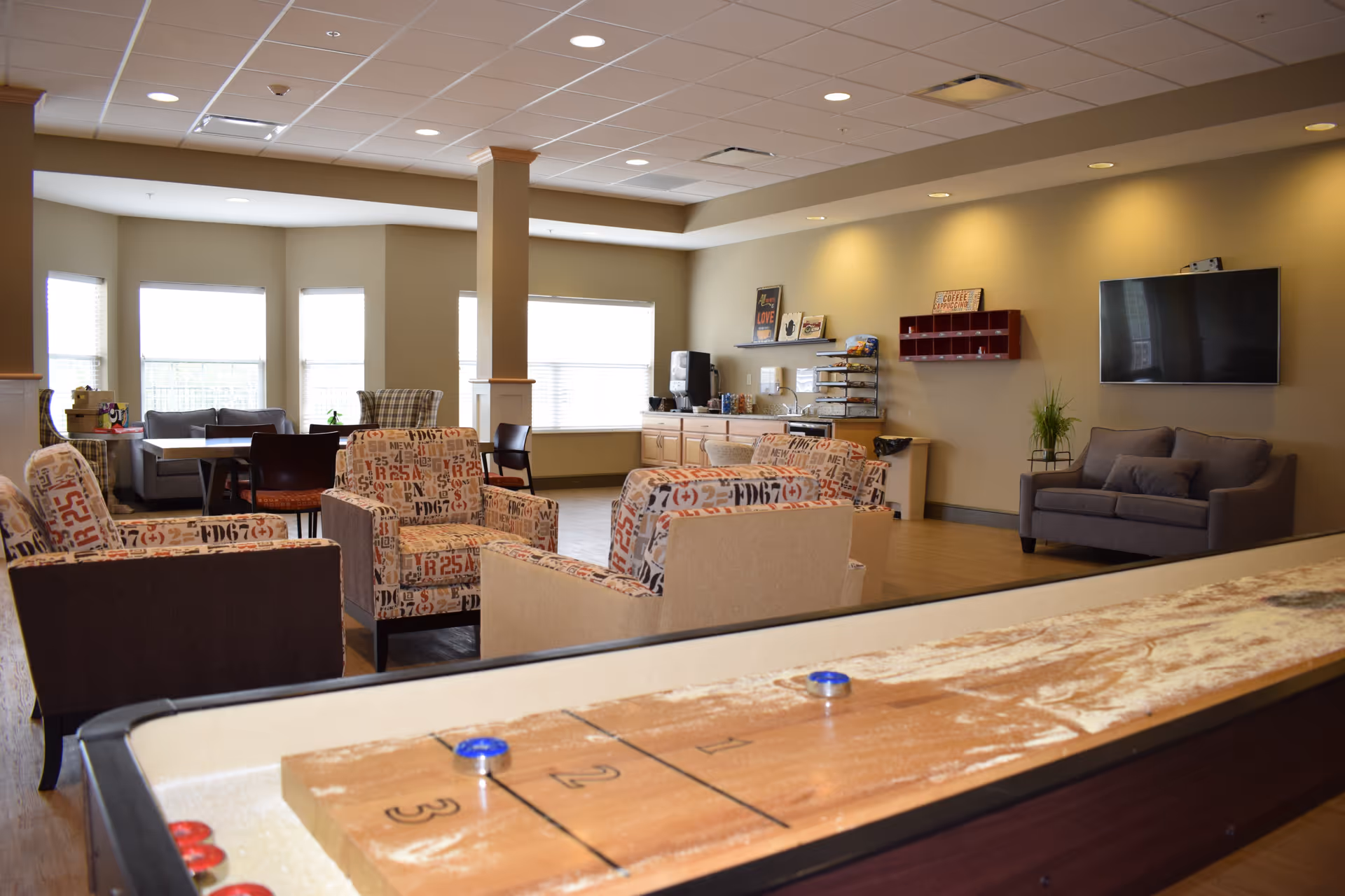 A spacious common area in a senior living facility with multiple patterned armchairs and a gray sofa arranged around a shuffleboard table. The room has large windows letting in natural light, a wall-mounted flat screen TV, a coffee station with supplies, and a few tables and chairs near the windows.