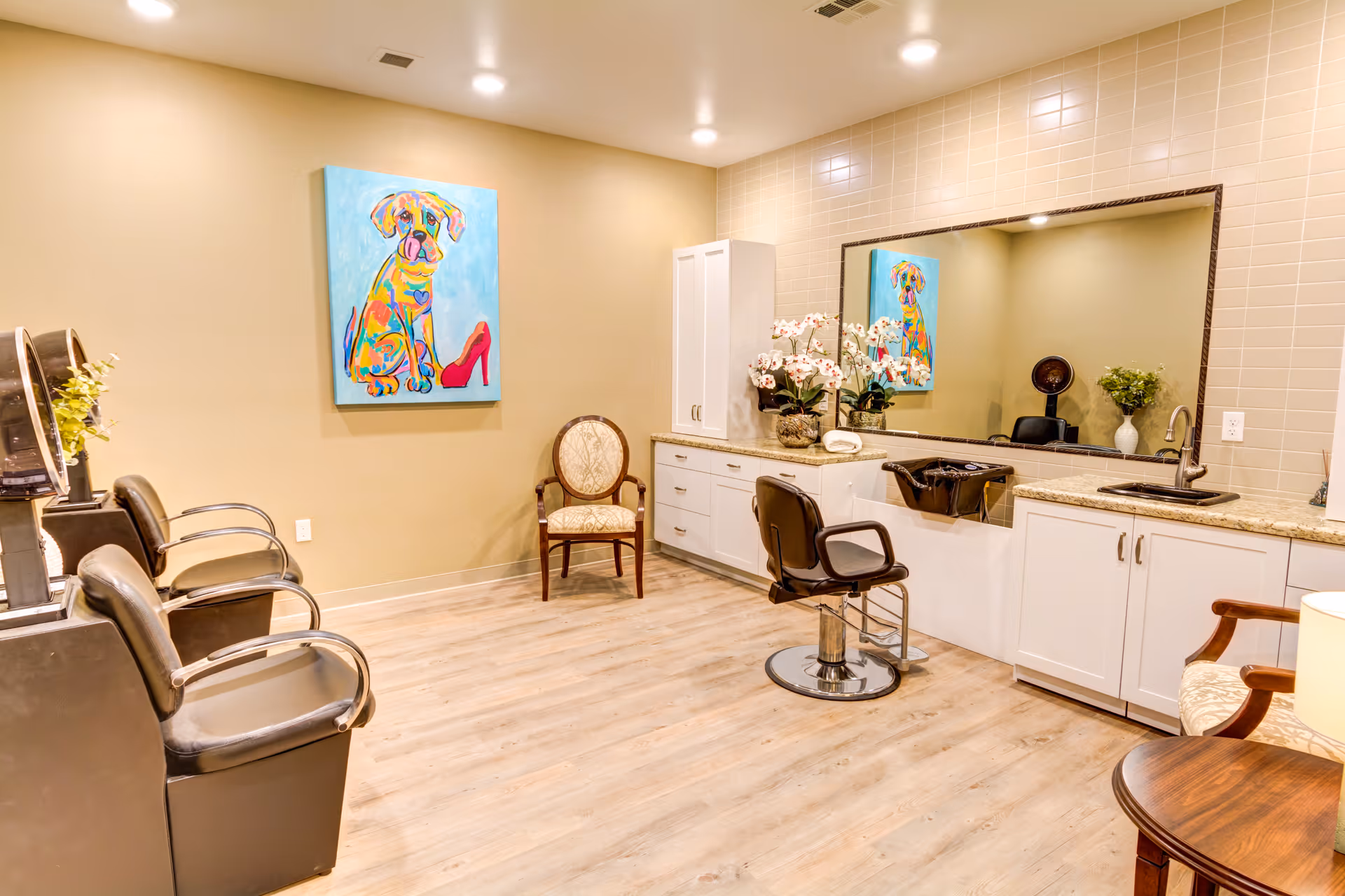 Interior of a hair salon area in a senior living facility with two black salon chairs facing hair dryers on the left, a single salon chair in front of a large mirror on the right, white cabinets and a sink beneath the mirror, a colorful painting of a dog with a red high heel on the wall, and two upholstered chairs with wooden arms.