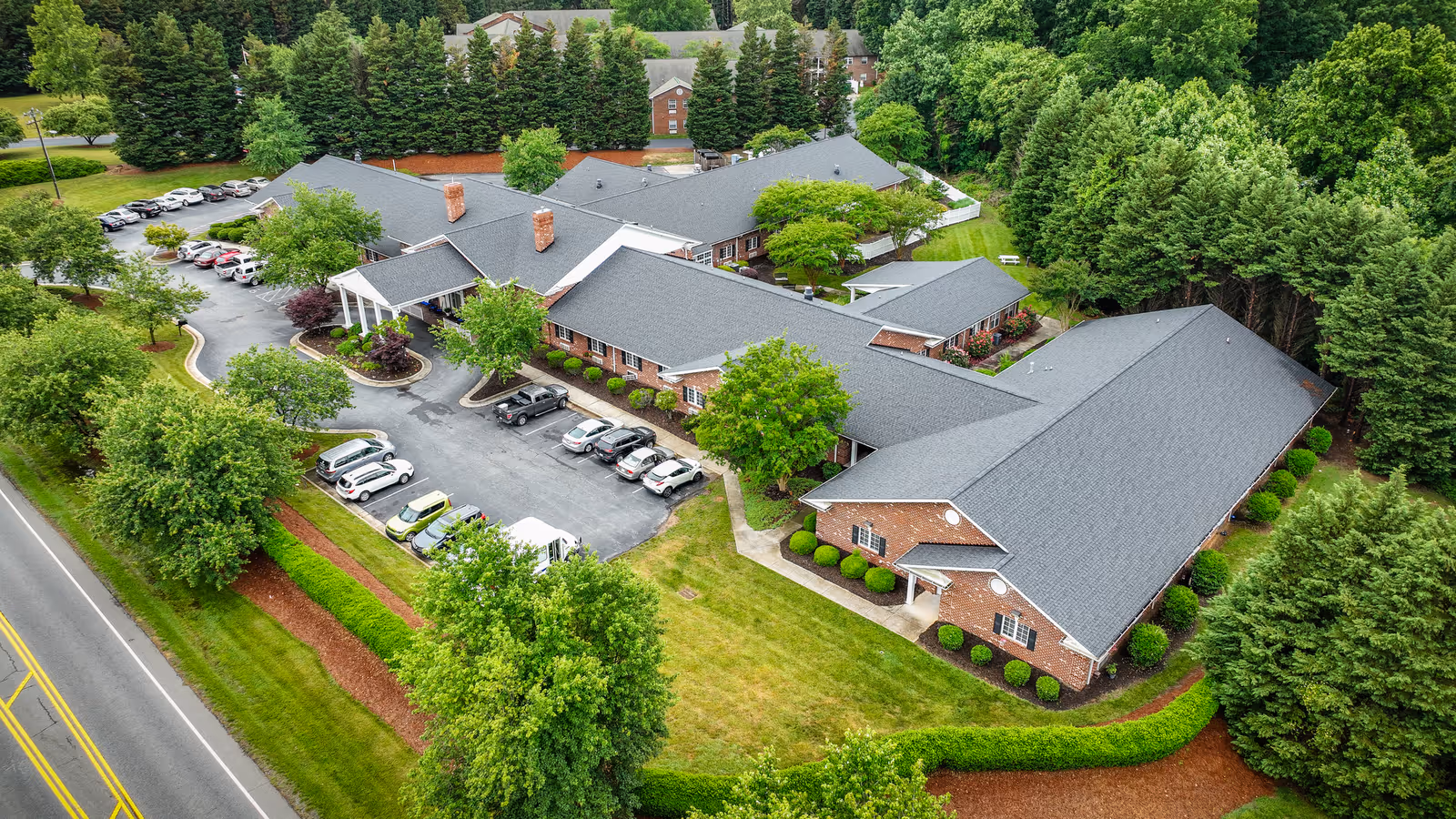 Aerial view of Kerner Ridge Assisted Living facility showing a large, single-story brick building with a gray roof surrounded by green trees and landscaped lawns. Several cars are parked in the parking lot adjacent to the building, and a road runs along the left side of the image.