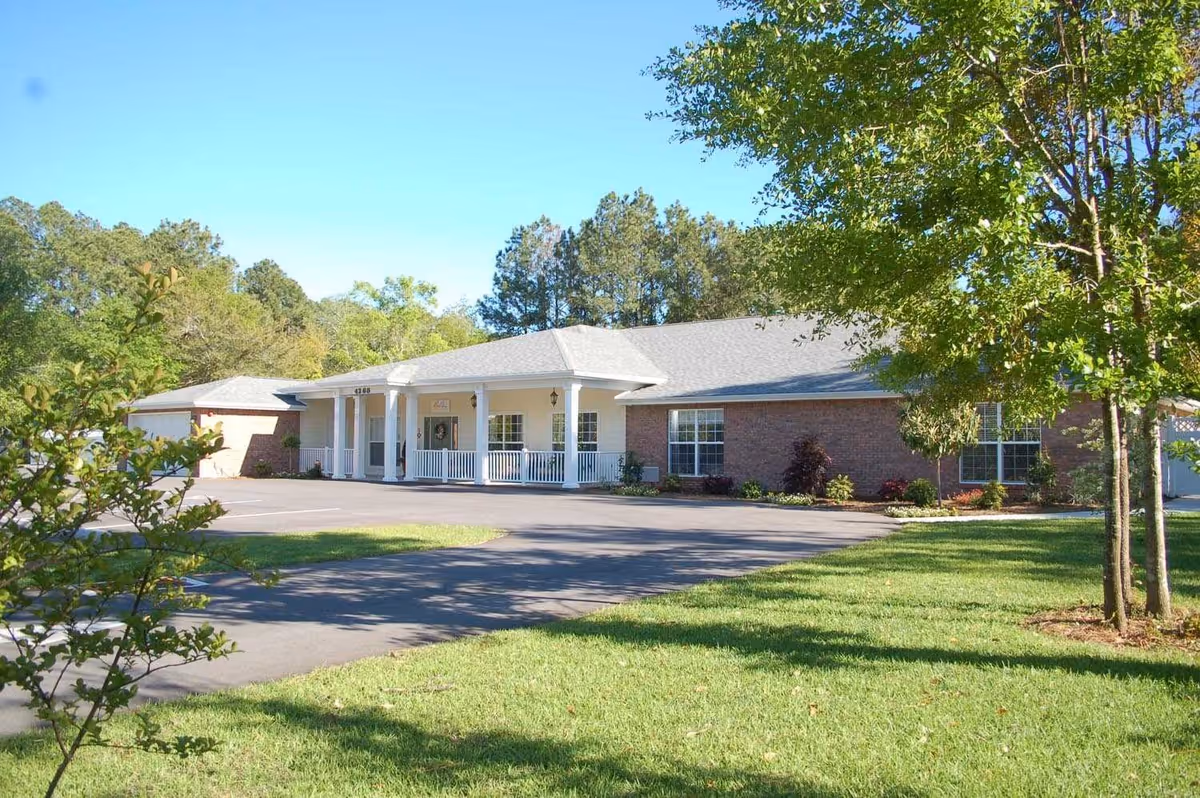 A single-story brick building with a white porch and columns, surrounded by green grass and trees under a clear blue sky. The building appears to be an assisted living facility with a paved driveway and parking area in front.
