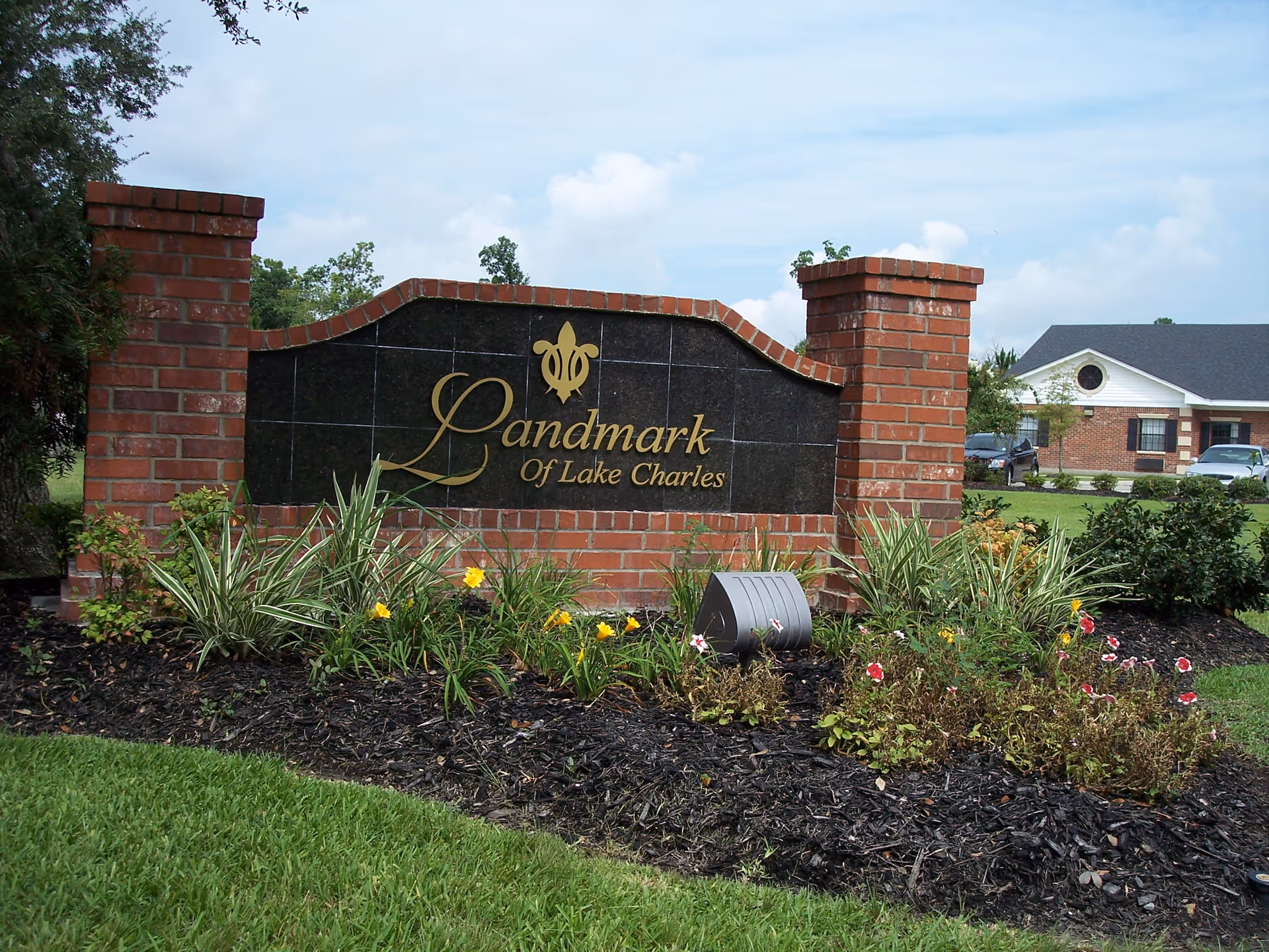 A brick and black stone sign with gold lettering that reads 'Landmark Of Lake Charles' surrounded by landscaped plants and flowers, with a building and parked cars visible in the background under a partly cloudy sky.