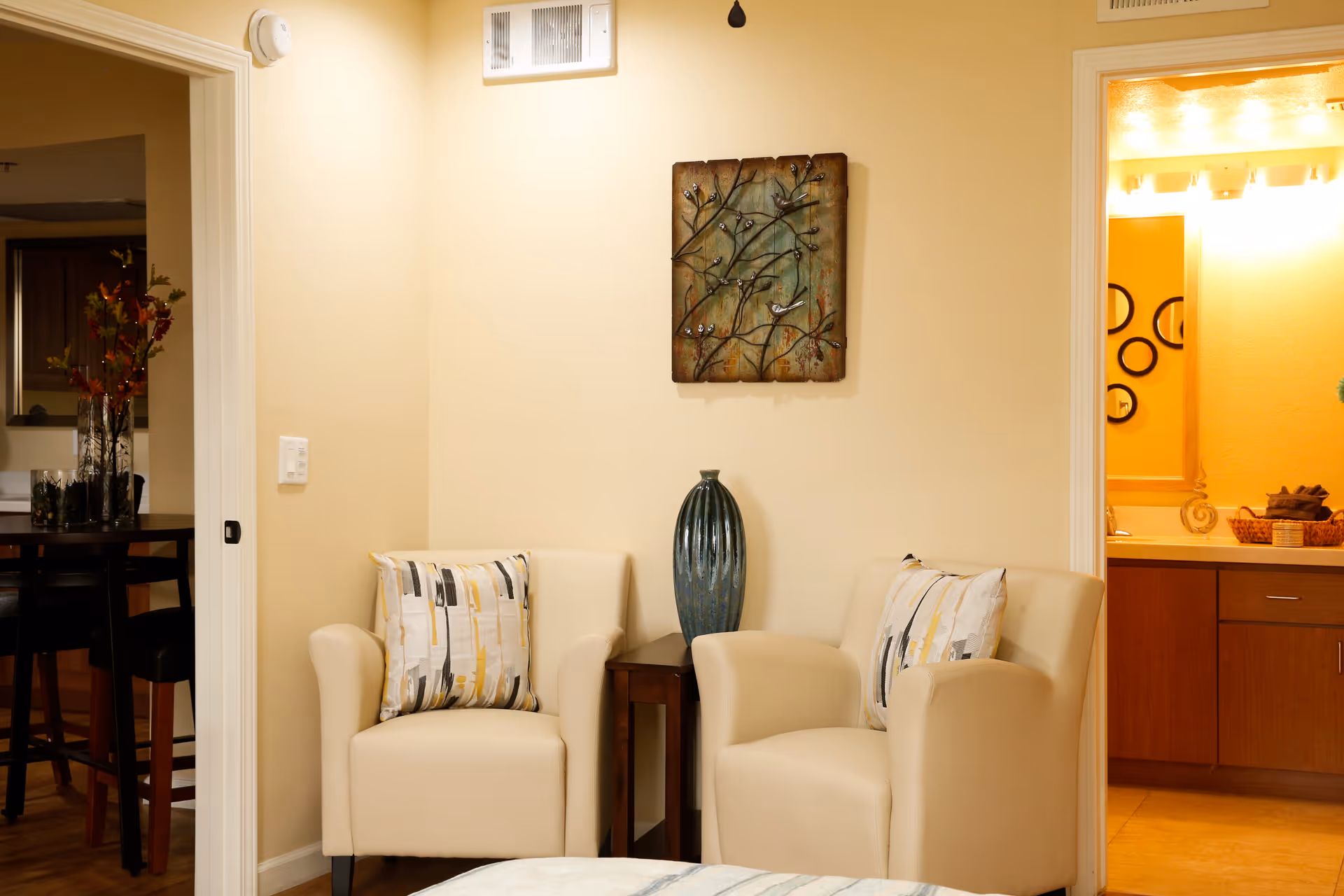 A cozy seating area with two beige armchairs, each with a decorative pillow, separated by a small wooden side table holding a tall blue vase. On the wall above the table is a metal wall art piece featuring branches and birds. To the left, there is an open doorway leading to a dining area with a table and chairs, and to the right, another open doorway reveals a bathroom with a sink and mirror.