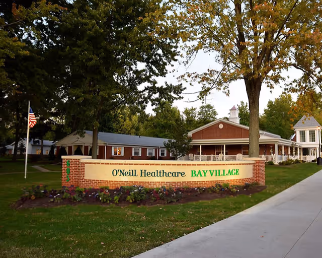 Brick entrance sign reading 'O'Neill Healthcare BAY VILLAGE' sits on a landscaped lawn in front of a single-story red-brick facility with trees.