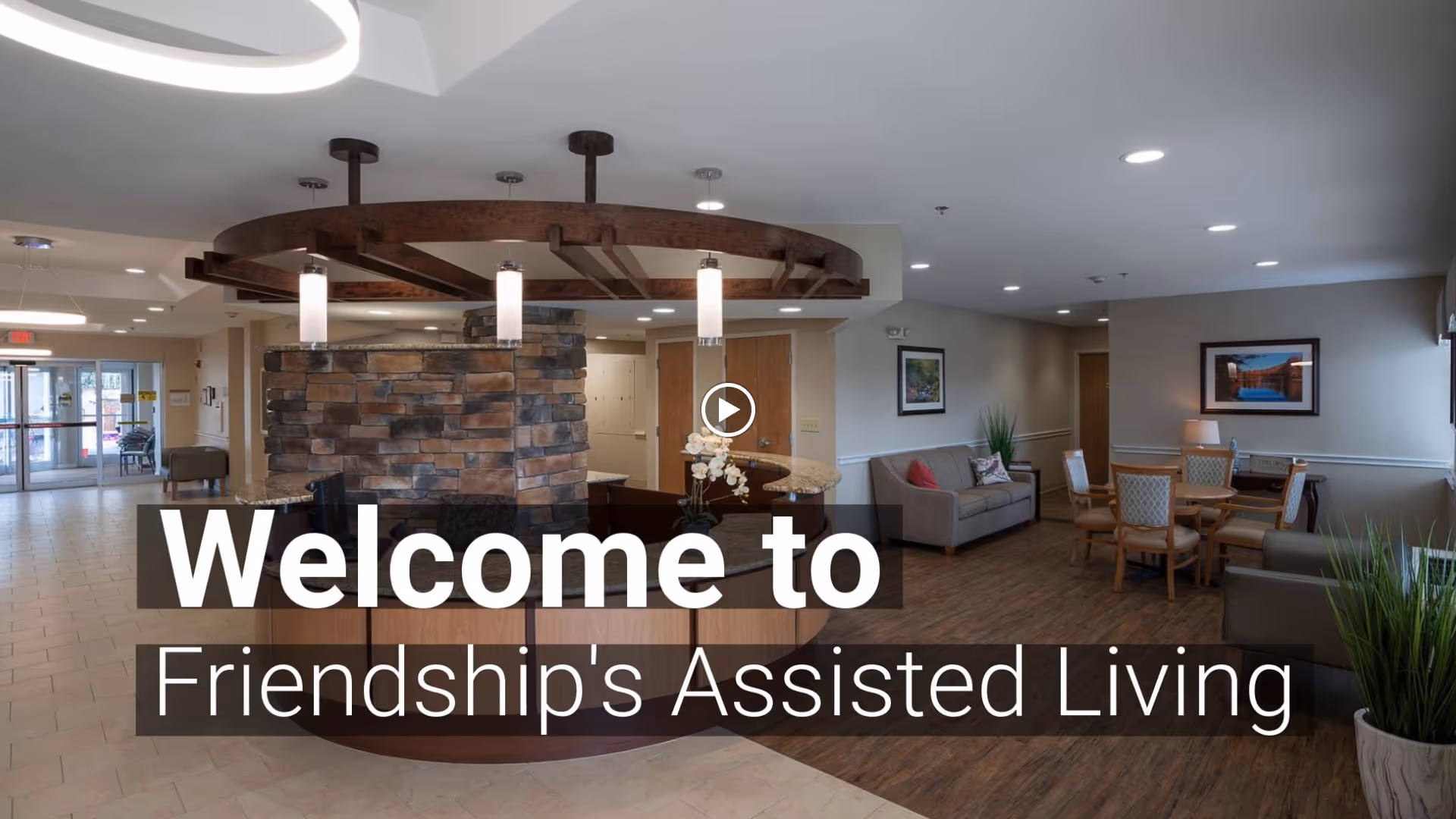 Interior view of the lobby area at Friendship Assisted Living featuring a circular reception desk with stone accents and hanging lights. The space includes seating areas with sofas and chairs, framed artwork on the walls, and plants, creating a welcoming atmosphere.
