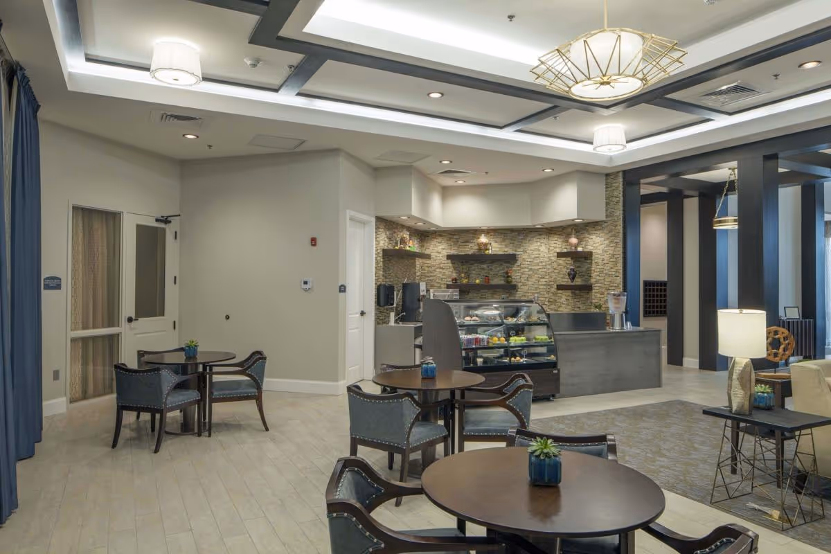 A modern and well-lit common area with round tables and chairs, a small counter with a glass display case containing food items, decorative shelves on a stone accent wall, and stylish ceiling lights. The space has light-colored flooring and a cozy seating area with a lamp and side table.