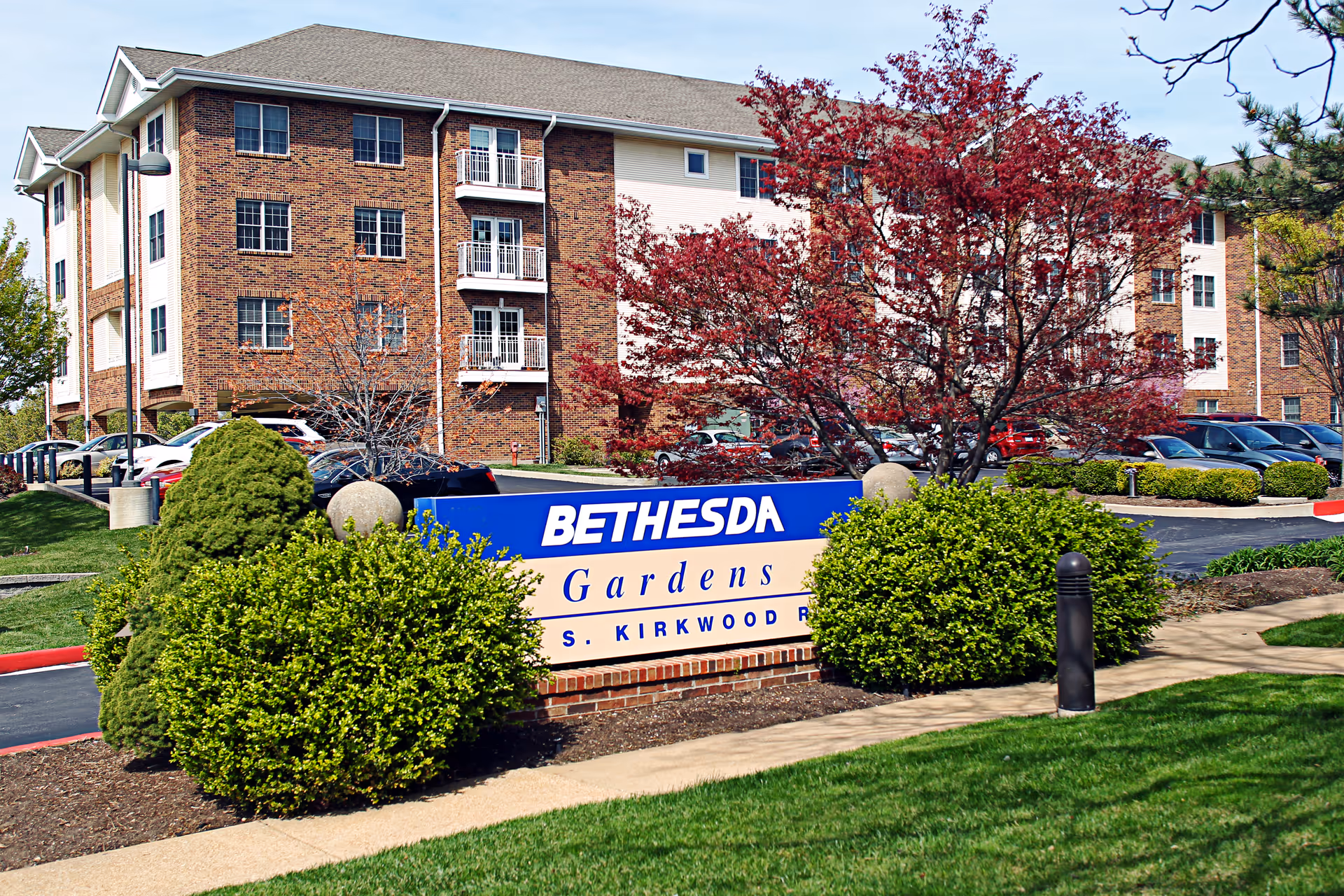 Exterior view of Bethesda Gardens senior living facility showing a brick building with multiple windows and balconies, surrounded by landscaped bushes, trees with red leaves, and a parking lot with cars. A blue and beige sign reading 'Bethesda Gardens S. Kirkwood Rd' is prominently displayed in front of the building.