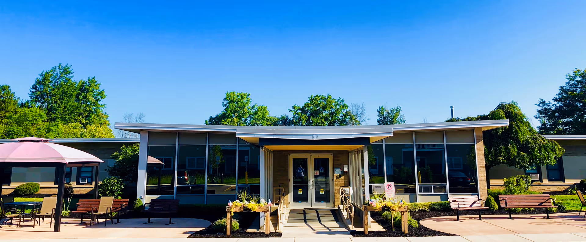 Front exterior view of Park Creek Senior Living Community building with large windows, benches, outdoor seating with umbrellas, and landscaped greenery under a clear blue sky.