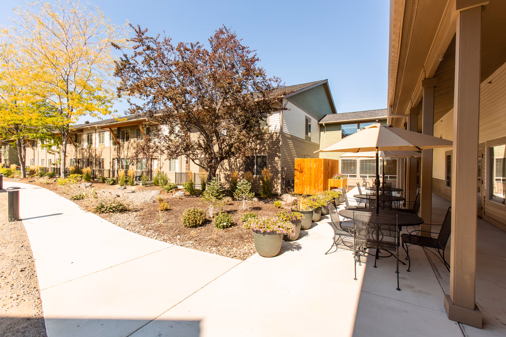 Outdoor courtyard with patio tables and umbrellas, potted plants, a walkway, and a two-story residential building.