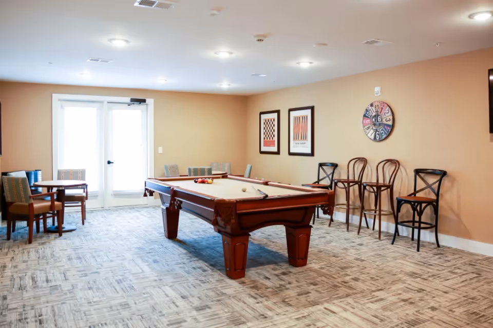 A well-lit game room with a pool table in the center, balls arranged on the table, and a cue stick resting on it. The room has beige walls with two framed game-themed pictures and a colorful clock on the wall. There are several chairs lined up against the wall and a small table with chairs near a set of double glass doors letting in natural light.