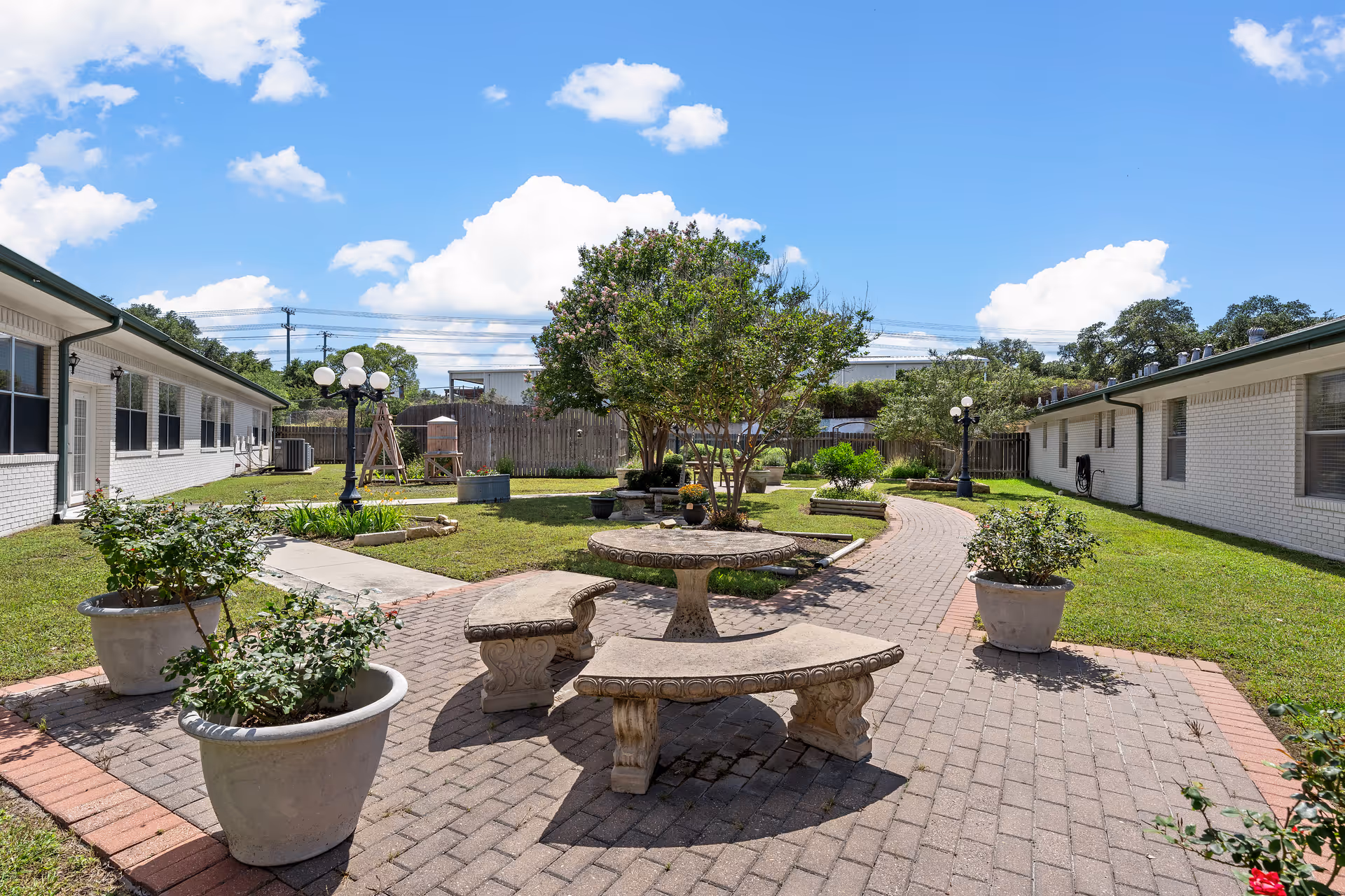 Sunny landscaped courtyard between single-story white brick buildings with paved pathways, stone benches, planters, and a central tree.