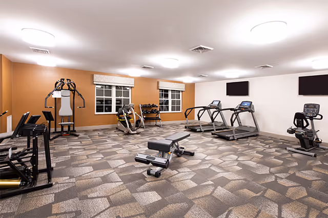 A spacious fitness room with various exercise equipment including treadmills, stationary bikes, weight machines, and free weights. The room has patterned carpet flooring, white walls with one orange accent wall, two windows with blinds, and ceiling lights providing bright illumination.