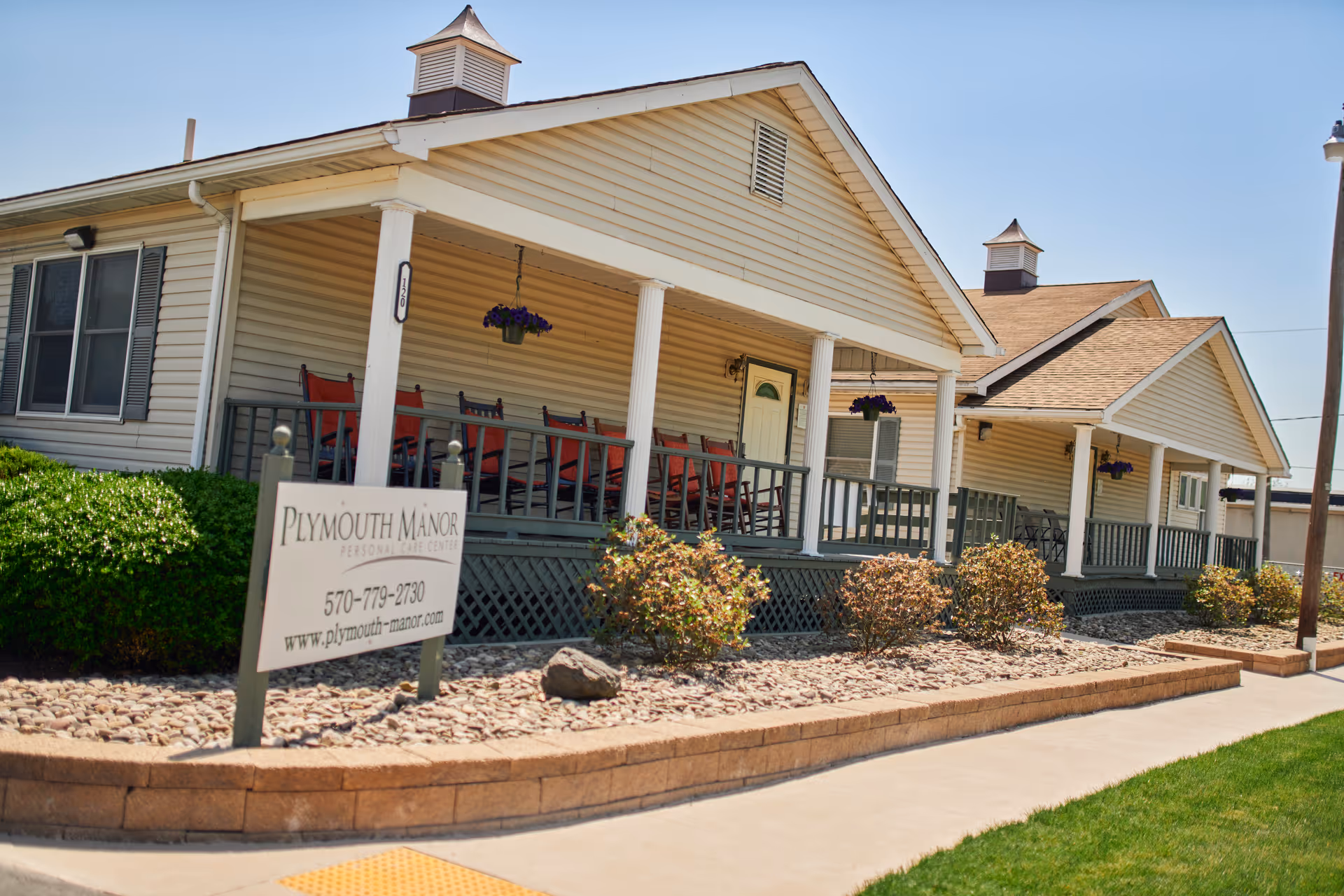 Exterior view of Plymouth Manor Personal Care Center showing a beige building with a covered porch featuring several red rocking chairs and hanging flower pots. There are bushes and landscaping rocks in front of the porch, and a sign with the facility's name, phone number, and website is visible near the sidewalk.