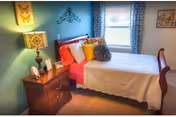 A cozy bedroom with a neatly made bed featuring white bedding and multiple colorful pillows. There is a wooden nightstand with a lamp, a small clock, and decorative items. The walls are painted blue and decorated with framed art and a wall-mounted butterfly decoration. A window with patterned curtains allows natural light into the room.
