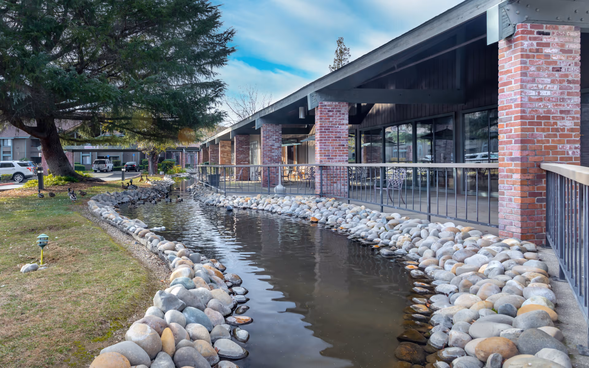 Brick-front senior living building with a covered patio and railing along a landscaped pond lined with river rocks.