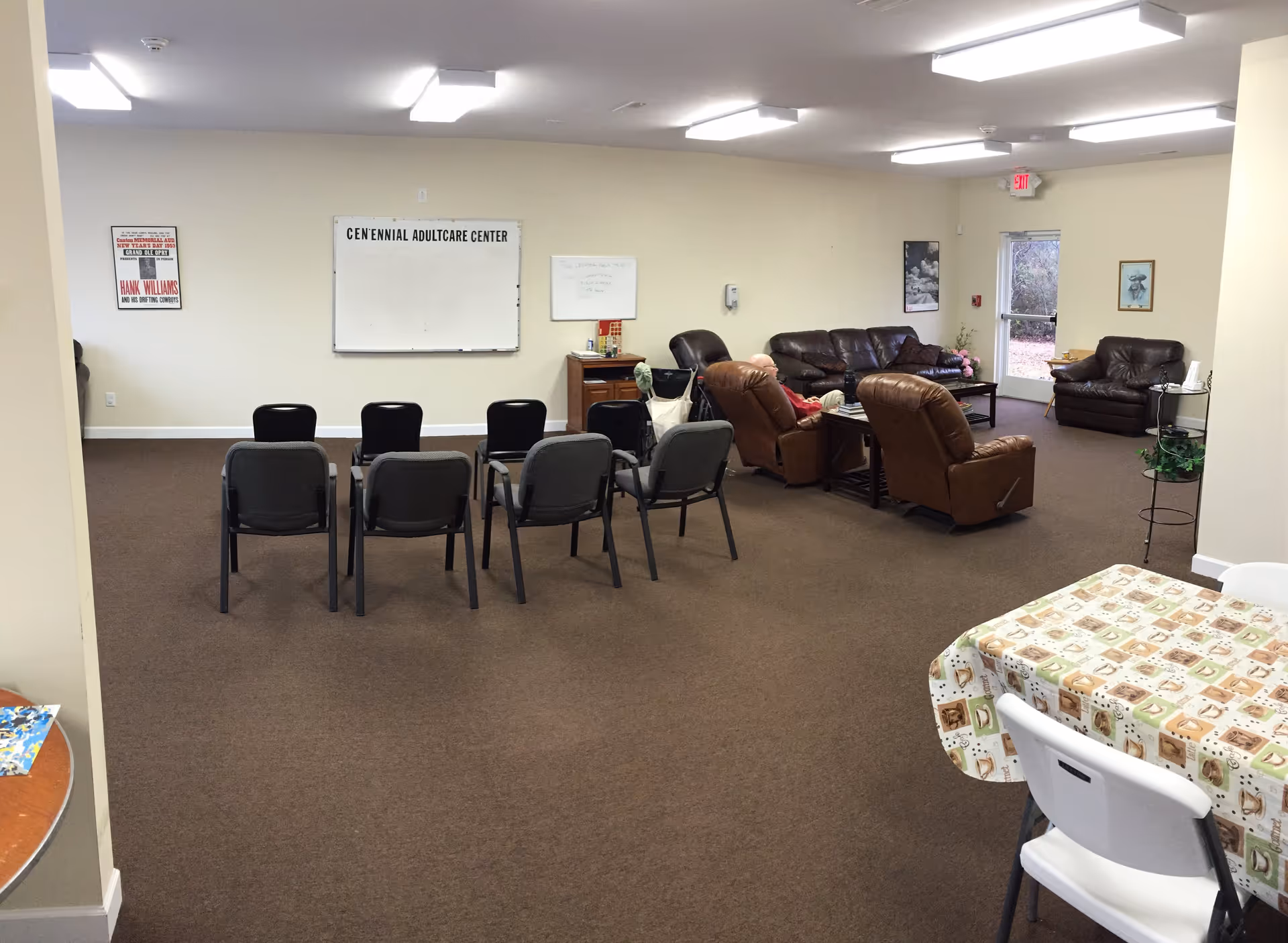 A spacious room in Centennial Adultcare Center with brown carpet and beige walls. The room contains a row of black chairs facing a whiteboard on the wall, several brown leather recliners and sofas arranged around a coffee table, and a table with a patterned tablecloth and white chairs in the foreground. There are framed pictures on the walls and a glass door leading outside.