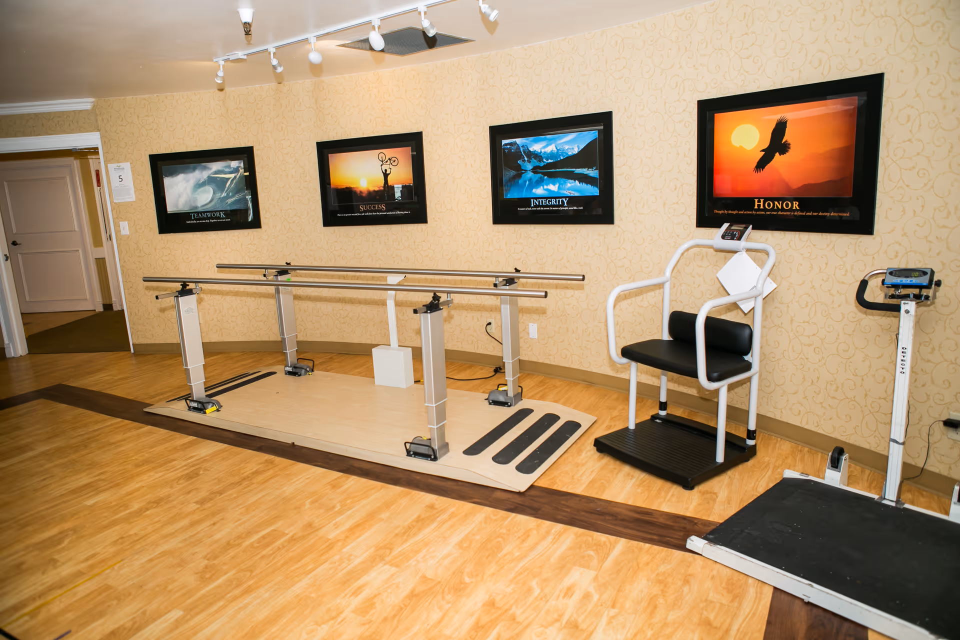 A physical therapy room with parallel walking bars, a therapy chair, scale, and framed motivational posters on the wall.