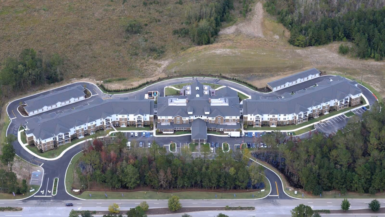 Aerial view of Okatie Pines Retirement Community showing a large, multi-wing building surrounded by parking lots, roads, and wooded areas.