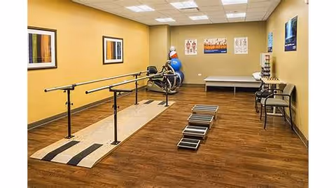 A physical therapy or rehabilitation room with parallel bars for walking exercises, a set of steps for stair training, exercise balls, chairs, and framed artwork on the walls. The room has wood flooring and beige walls with ceiling lights.