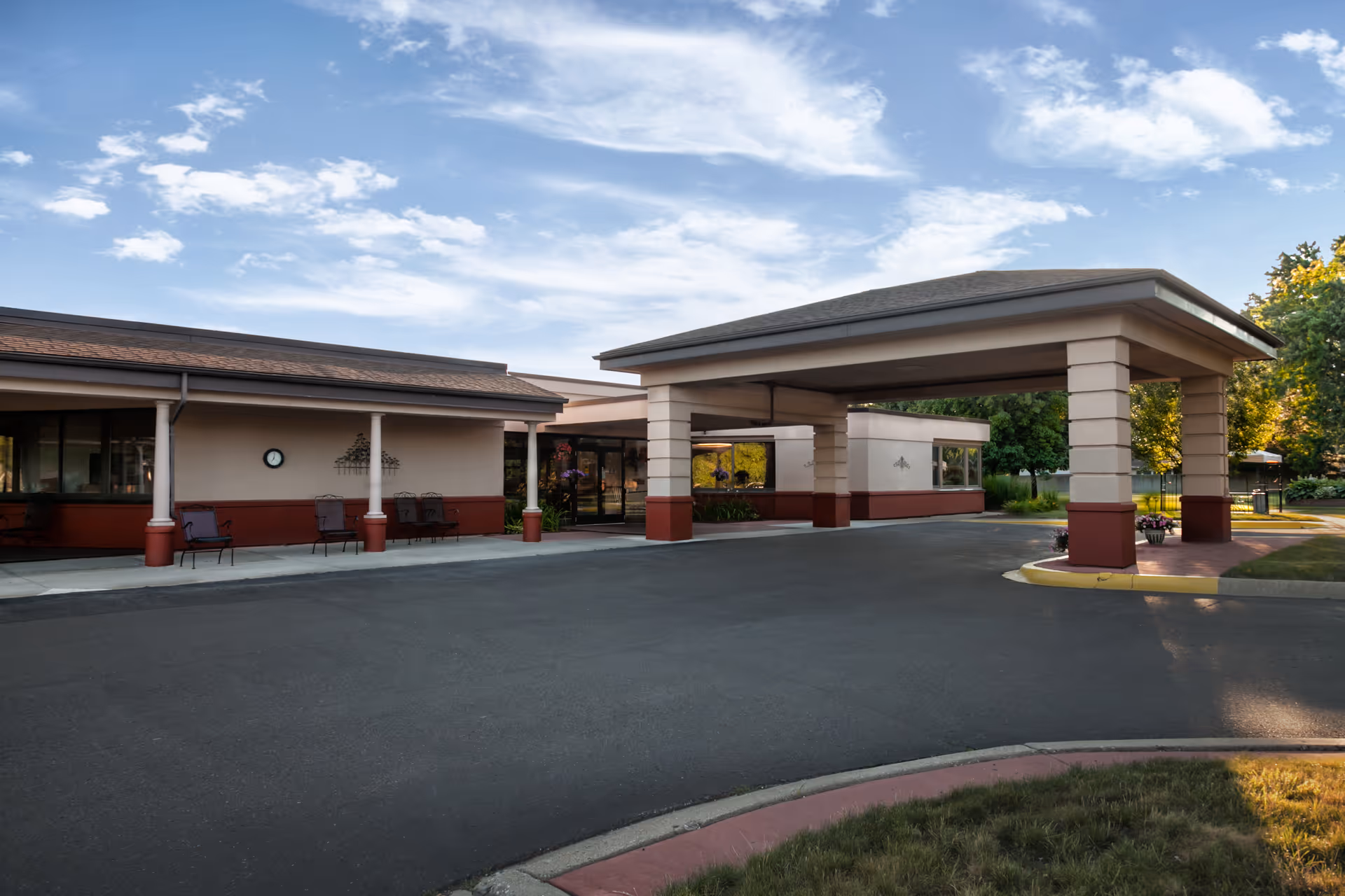 Exterior view of Autumn Woods Health Care Facility showing the entrance with a covered drop-off area, surrounded by trees and a clear sky.