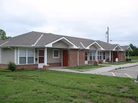 Row of single-story brick senior living buildings with entrances, windows, and a manicured lawn.