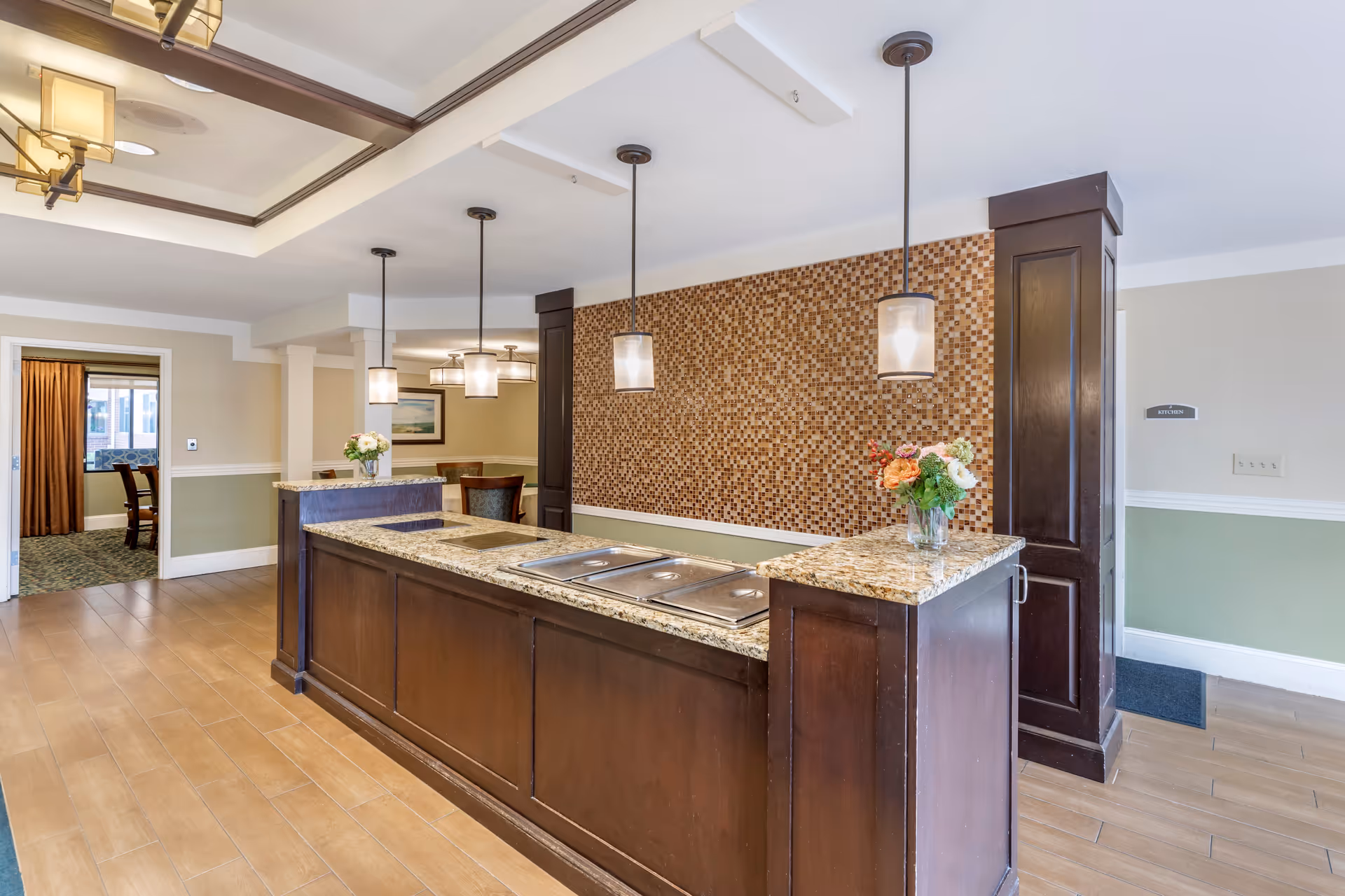 Interior view of a senior living facility featuring a long granite countertop with dark wood cabinetry and three pendant lights hanging above. The back wall has a mosaic tile design. There is a vase with flowers on the counter. In the background, there is a dining area with chairs and a framed picture on the wall.