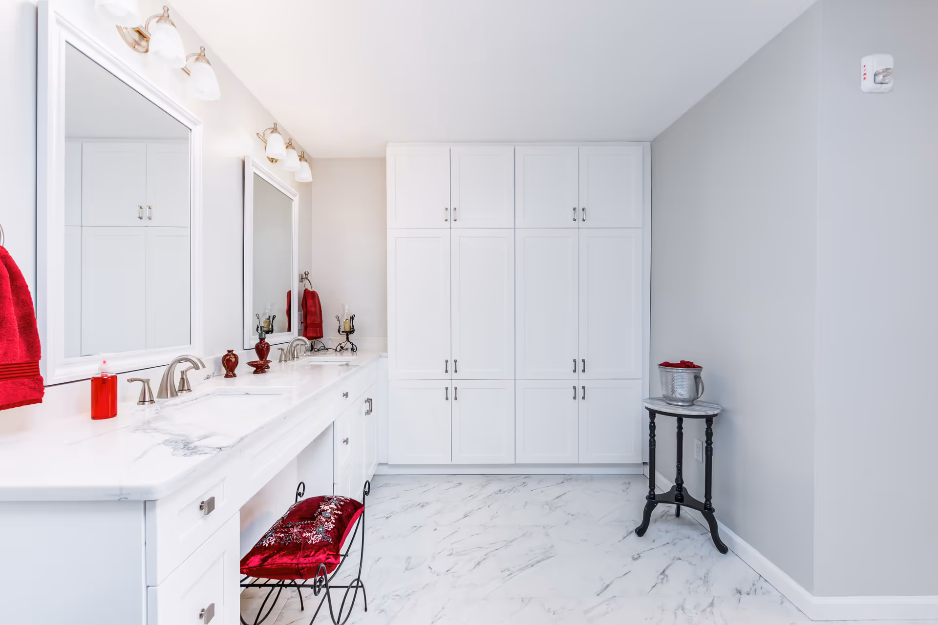 Bright and spacious bathroom with white marble countertops, double sinks, large mirrors, white cabinetry, and a red cushioned stool. The floor is white with gray marble patterns, and there are red towels and decorative items adding color. A small round table with a silver bucket and red cloth is placed against a gray wall.