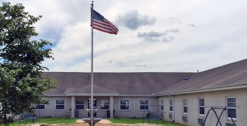 Exterior view of a single-story healthcare facility building with a large American flag on a flagpole in the center foreground. The building has multiple windows and a sloped roof, with a tree on the left side and swings on the right side in a grassy area under a partly cloudy sky.