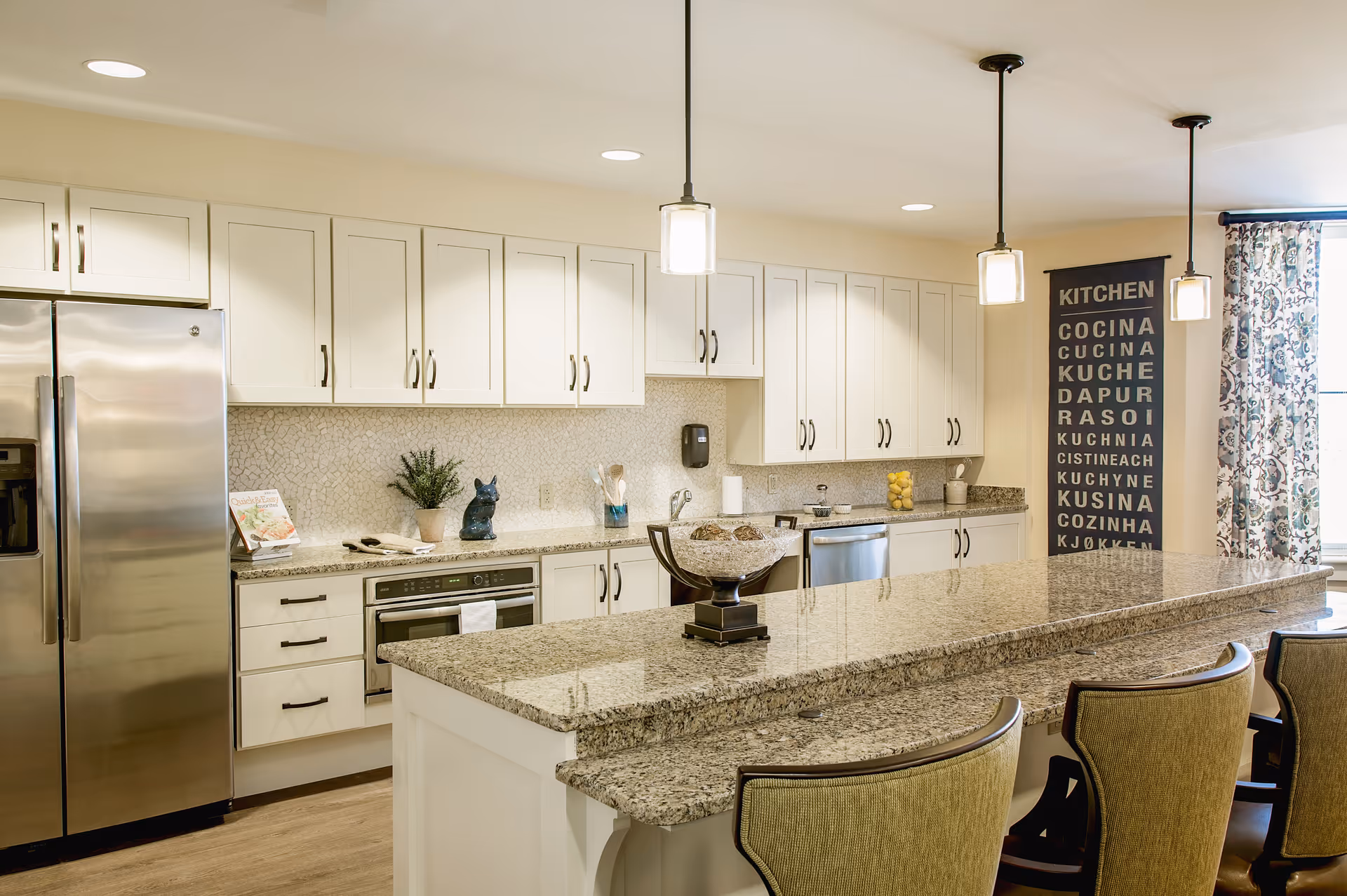 A modern kitchen with white cabinets, stainless steel refrigerator, built-in oven, and granite countertops. There is a large kitchen island with three upholstered chairs. Pendant lights hang above the island, and a decorative sign with the word 'KITCHEN' in multiple languages is displayed on the wall near a window with patterned curtains.