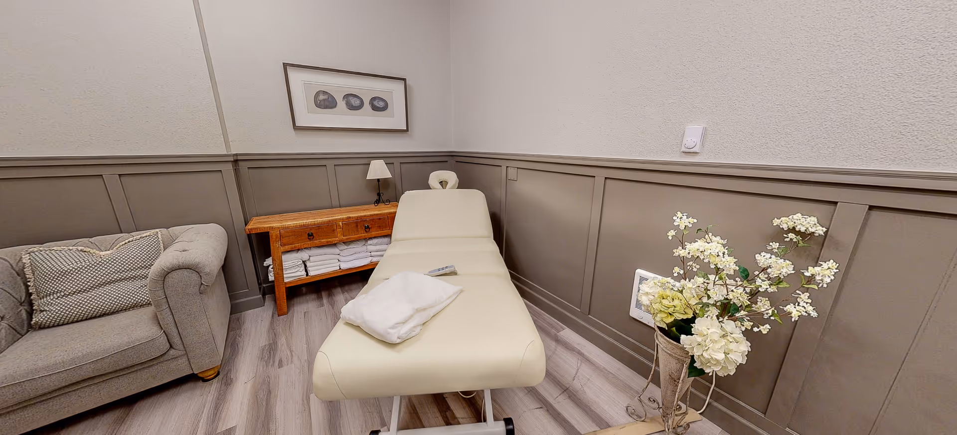 A small room with a beige massage table covered with a white towel, a gray upholstered armchair with a textured pillow, a wooden side table with folded white towels and a small lamp, a framed artwork on the wall, and a vase with white flowers on the floor.