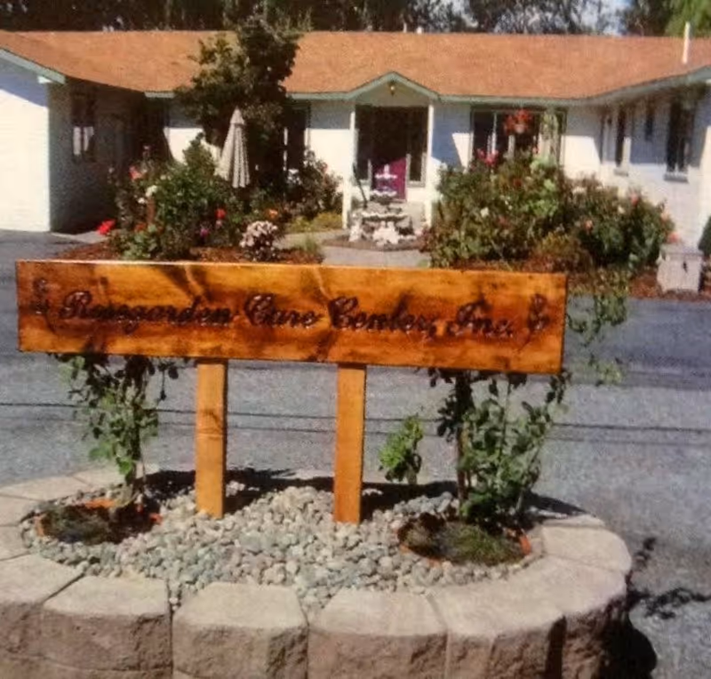 A wooden sign reading 'Rosegarden Care Center, Inc.' is mounted on two posts surrounded by small rocks and plants. Behind the sign, there is a single-story building with a brown roof, white walls, and a small garden area with bushes and flowers in front of the entrance.