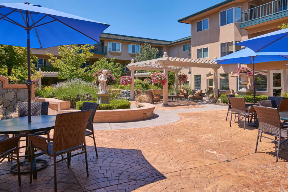 Outdoor courtyard area at The Springs at Anna Maria featuring round tables with wicker chairs and blue umbrellas, a pergola with hanging flower baskets, a statue, and surrounding greenery with a multi-story building in the background.