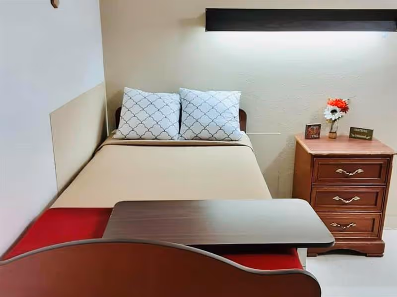 A simple single bedroom with a made bed, two patterned pillows, an overbed table, and a wooden nightstand topped with a small vase of flowers.
