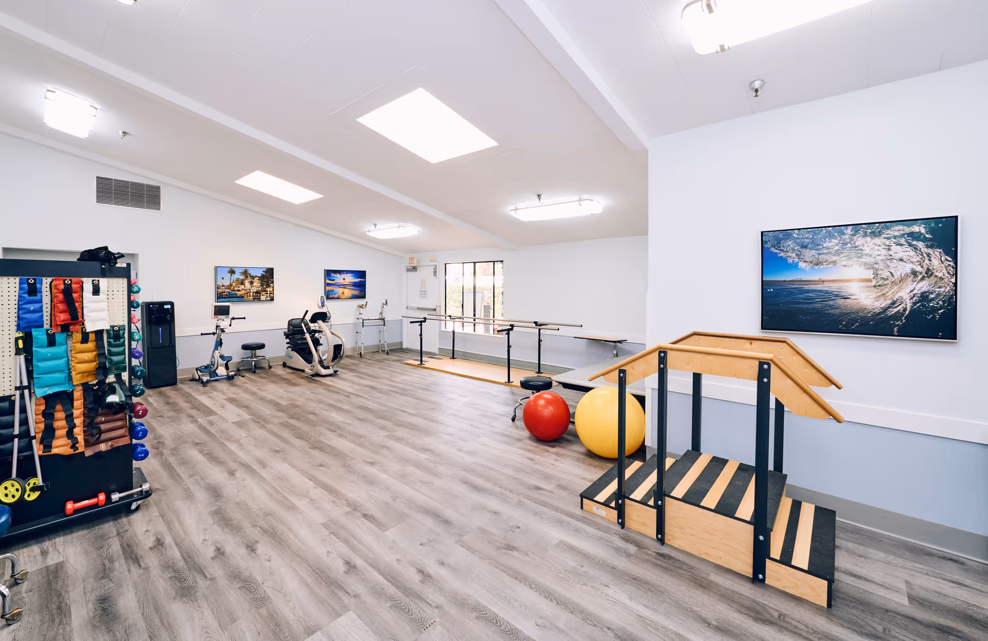 A spacious rehabilitation or physical therapy room with exercise equipment including stationary bikes, therapy balls, hand weights, resistance bands, and a wooden step platform with handrails. The room has light wood flooring, white walls, ceiling lights, and framed pictures of ocean scenes on the walls.
