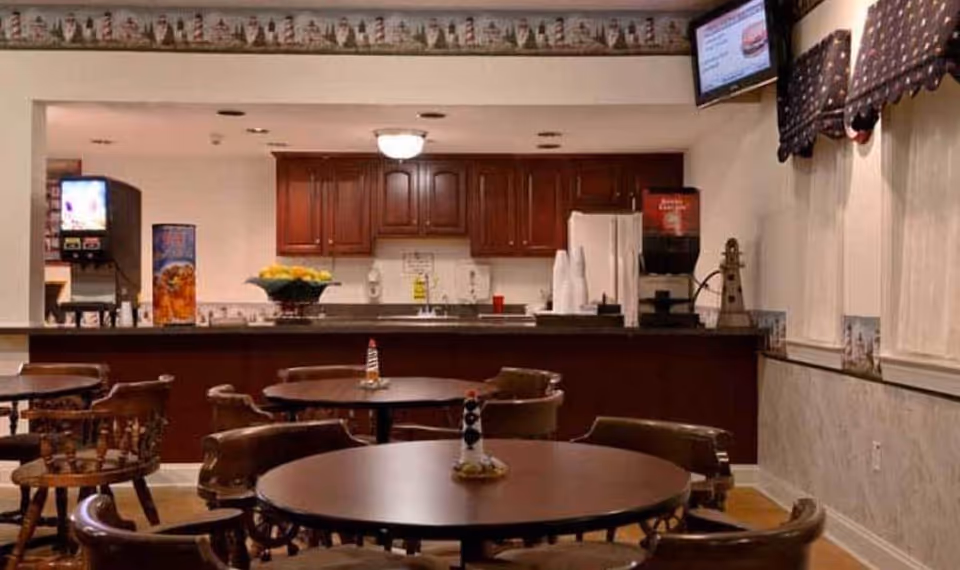 Interior view of a dining area with round wooden tables and chairs. In the background, there is a kitchen counter with a sink, a refrigerator, a beverage dispenser, and cabinets. The walls have a decorative border near the ceiling and windows with valances. A TV screen is mounted on the upper right wall.