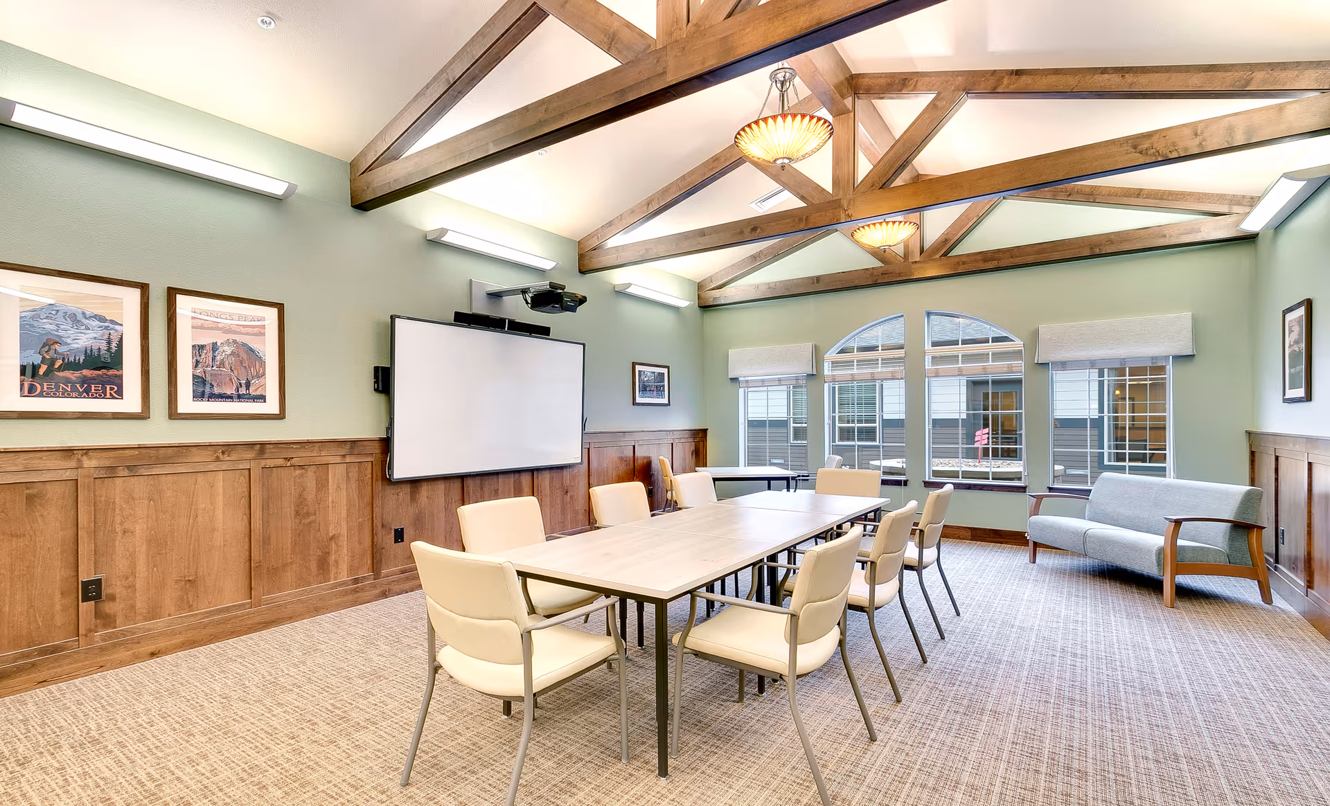 A well-lit meeting or conference room with a long rectangular table surrounded by beige chairs. The room features wooden beams on the ceiling, green walls with wood paneling, framed artwork, a large whiteboard or screen mounted on the wall, and three large windows with beige valances. There is also a gray upholstered bench along one wall.
