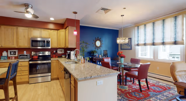A modern kitchen and dining area in a senior living facility. The kitchen features wooden cabinets, stainless steel appliances including a microwave and oven, a granite countertop island with a sink, and bar stools. The dining area has a round glass table with red chairs, a colorful patterned rug, and large windows with blue and white curtains allowing natural light to fill the room. The walls are painted in warm tones with one accent wall in blue, decorated with a mirror and artwork.