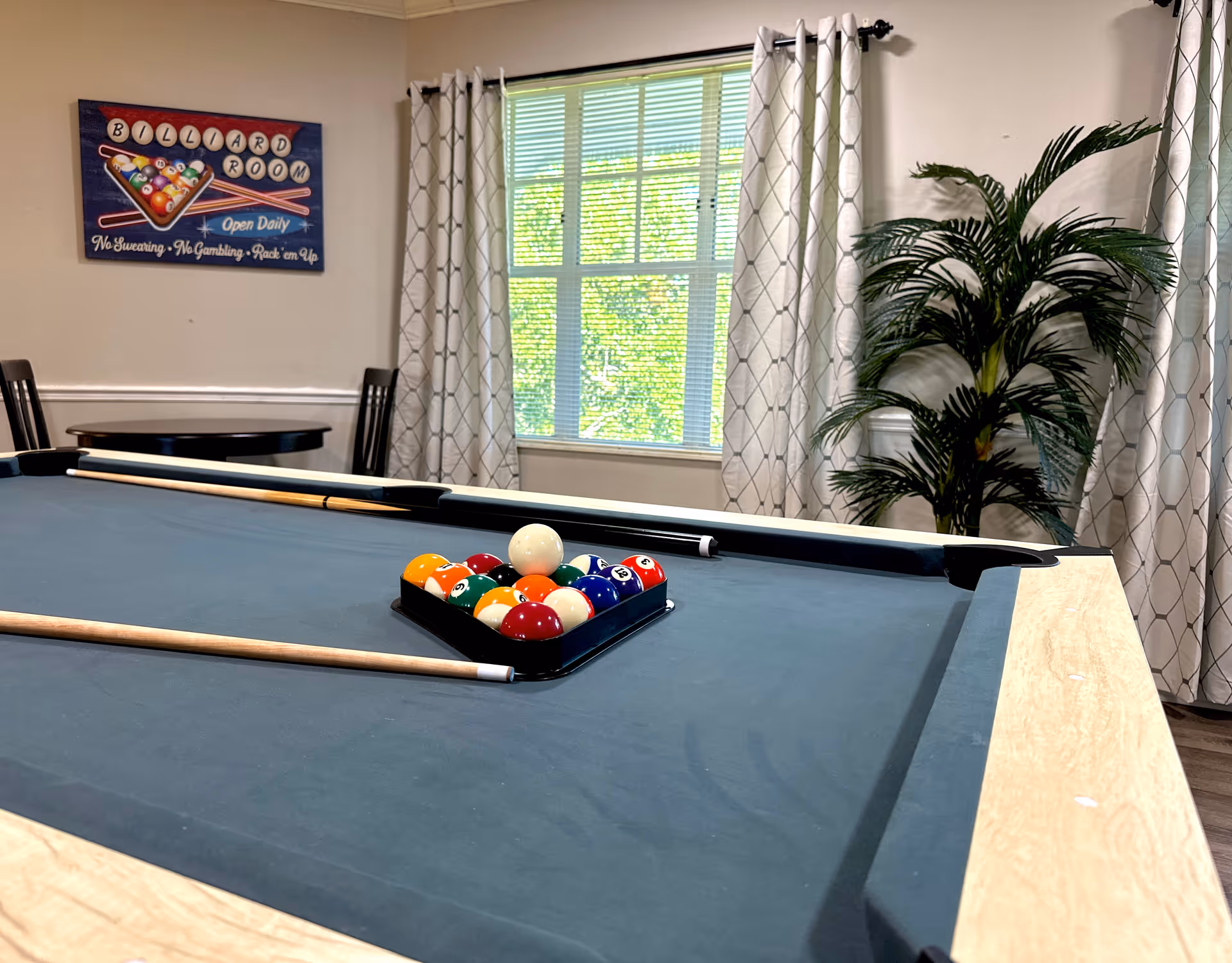 A billiards table with racked balls and cues in a bright interior room with a window, curtains, a potted plant, and a wall sign.