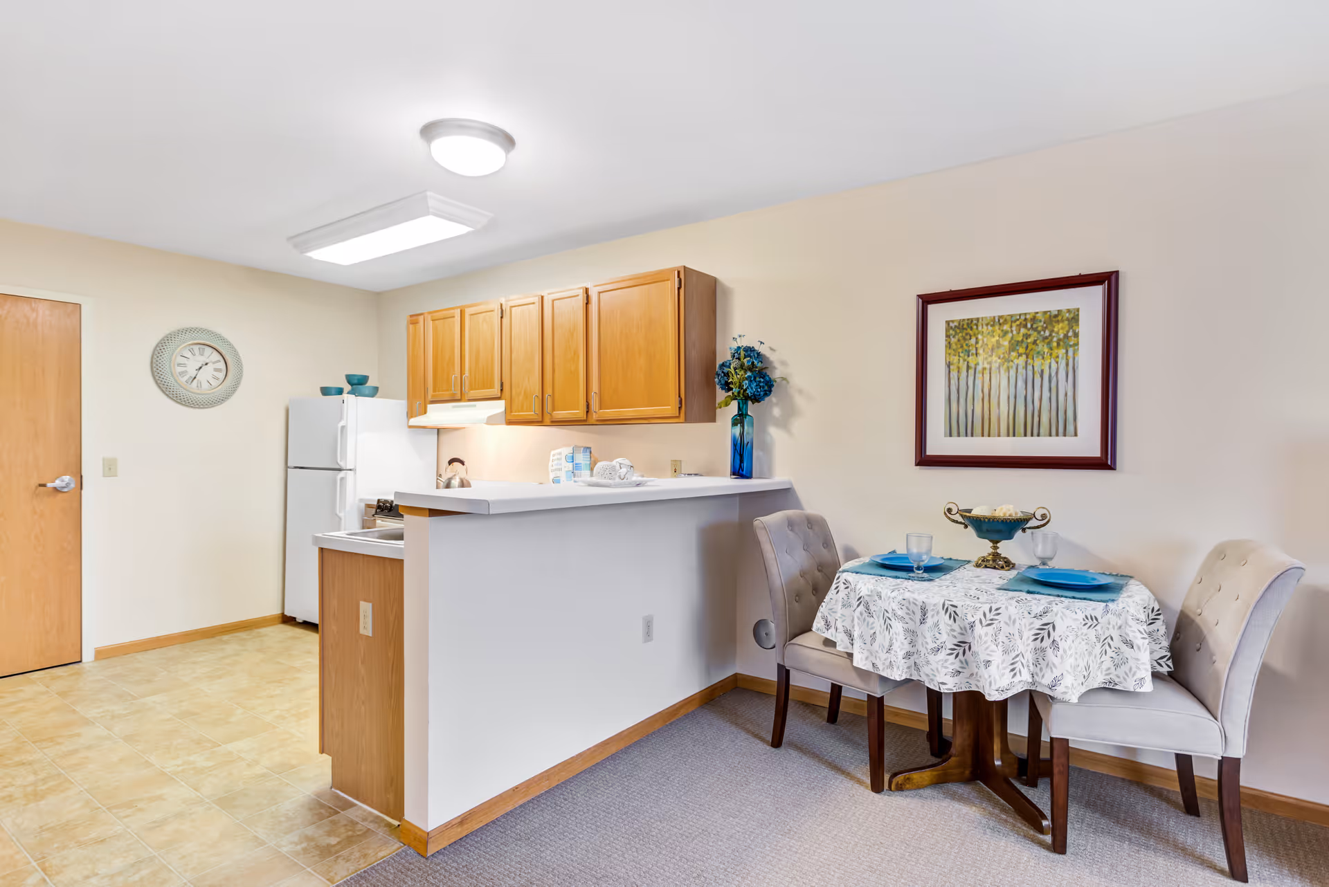 A small dining area and kitchen in a senior living facility. The dining area has a round table covered with a white patterned tablecloth, set with two blue plates, glasses, and a decorative centerpiece. Two beige upholstered chairs are placed around the table. The kitchen features wooden cabinets, a white refrigerator, and a countertop with a small overhang. A wall clock is mounted near a wooden door, and a framed picture of trees hangs on the wall above the dining table.