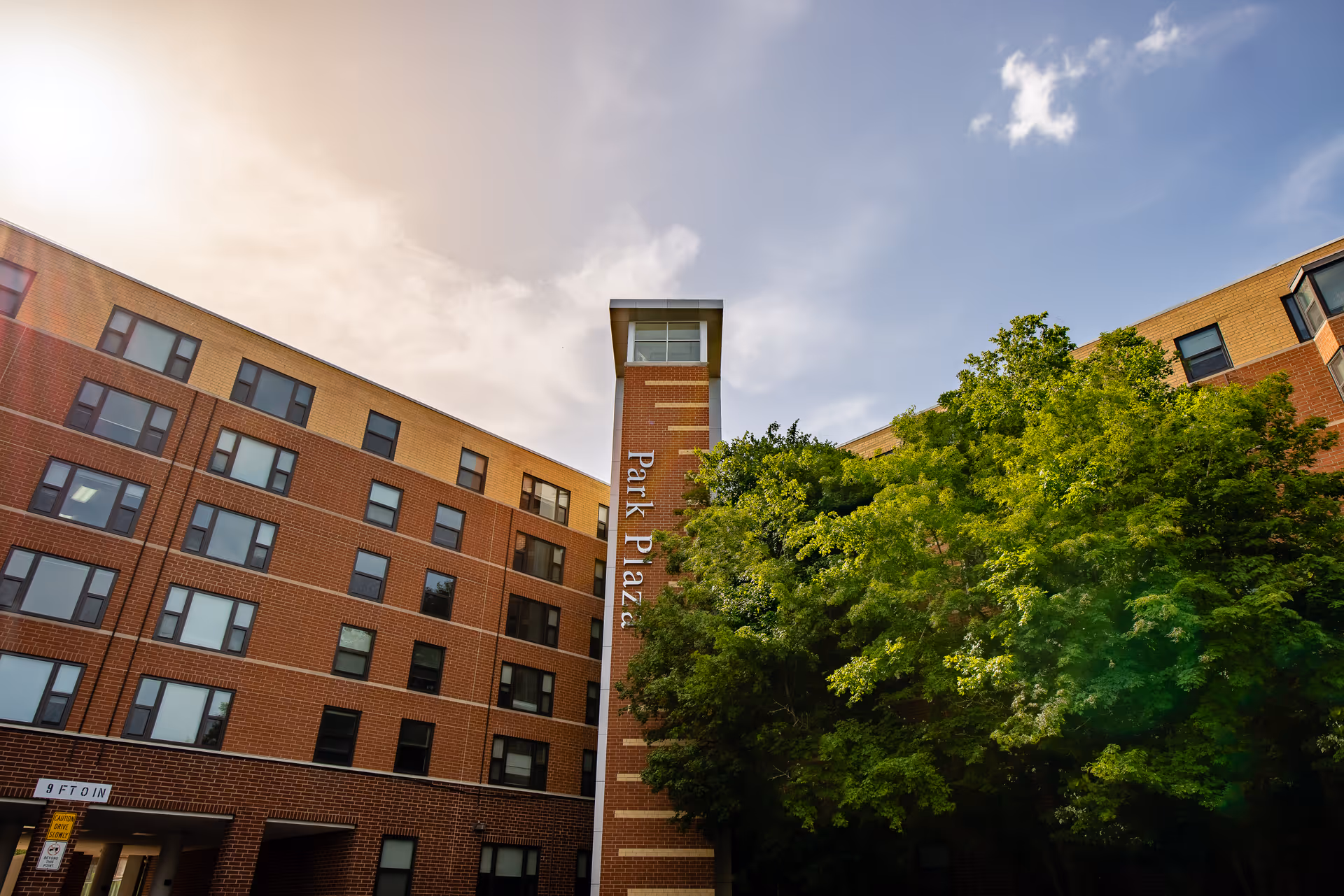 Exterior view of Park Plaza Retirement Center, a multi-story brick building with numerous windows. The building has a central tower with the name 'Park Plaza' displayed vertically. There are green trees in front of the building and a partly cloudy sky above.