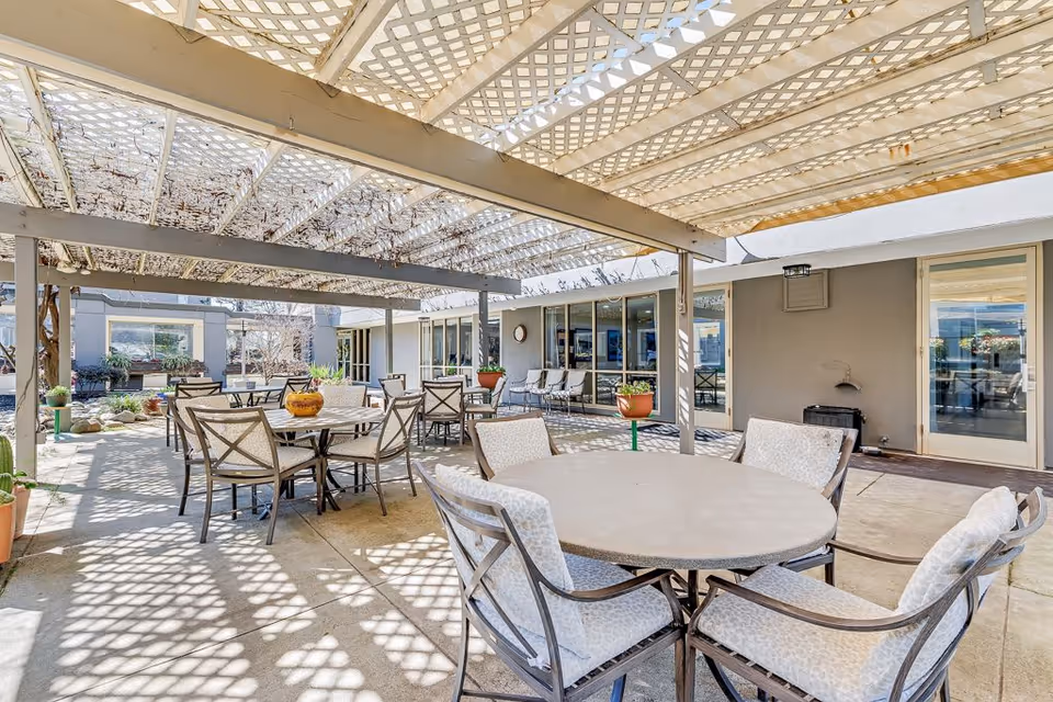 Outdoor patio area at a senior living facility with multiple round tables and cushioned chairs under a lattice pergola casting patterned shadows on the ground. Potted plants and large windows of the building are visible in the background.