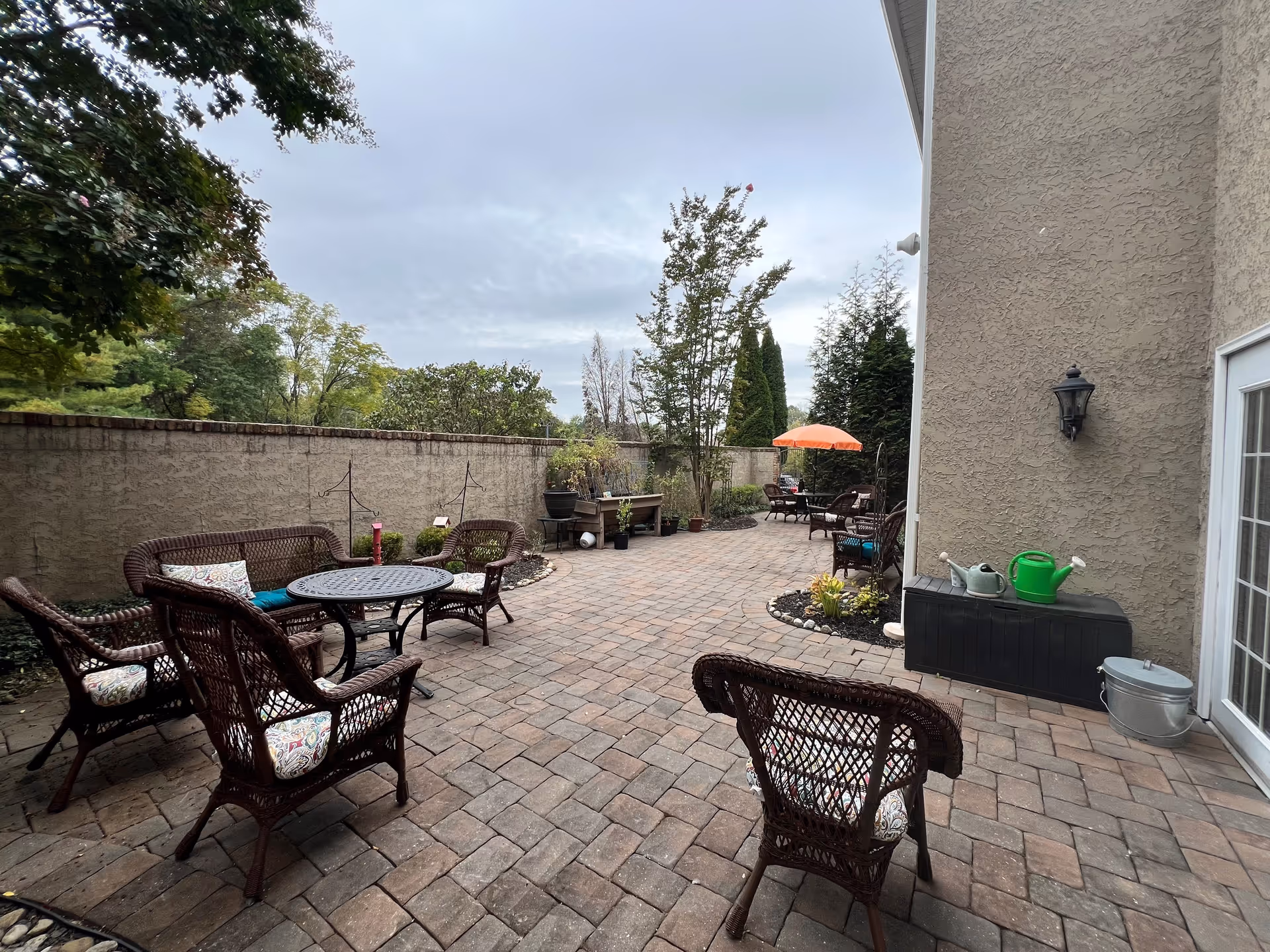Outdoor patio area with brick flooring, several wicker chairs with cushions, a round metal table, potted plants, and an orange umbrella in the background. The area is enclosed by a stucco wall and adjacent to a building with a light fixture and a door.