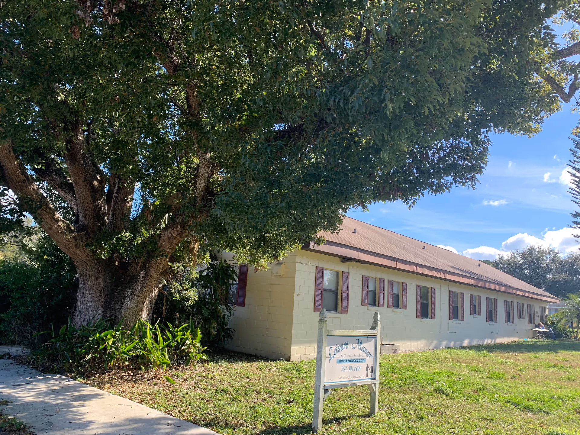 Exterior view of a single-story senior living building with a large tree and sign in front.
