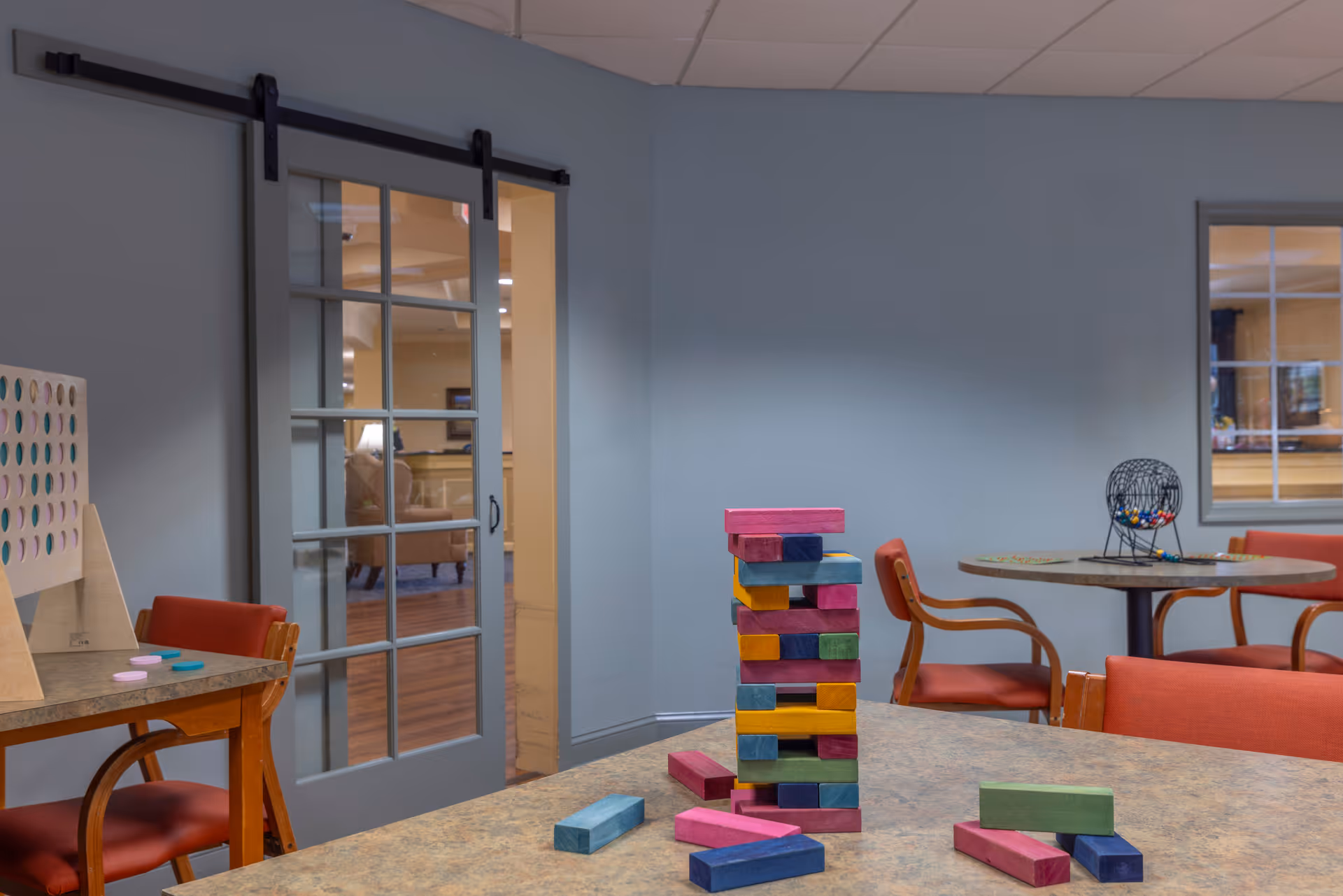 A common area in an assisted living facility with a table featuring a colorful wooden block stacking game and a bingo cage on another table. The room has red chairs, a sliding glass door, and a light blue wall.