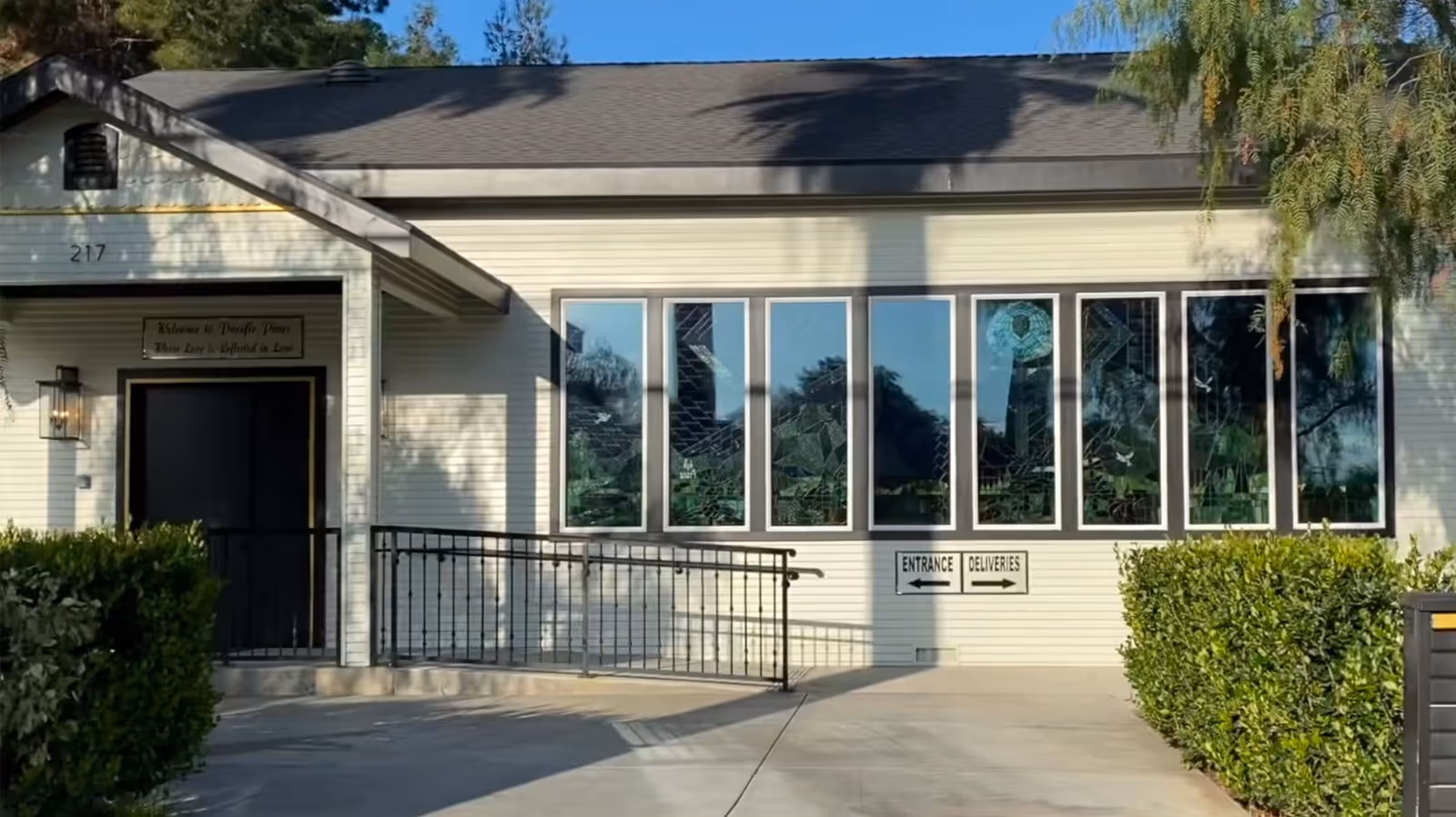 Exterior view of Pacific Pines Redlands building with a ramp leading to a black door under a small porch. The building has white siding, a dark roof, and several tall windows with decorative stained glass. There are bushes on either side of the walkway and signs indicating entrance and deliveries.