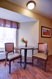 A small round wooden table with a vase of flowers on top, flanked by two wooden chairs with striped cushions, set near a window with blinds and a purple valance in a warmly lit room with wooden flooring and a framed picture on the wall.