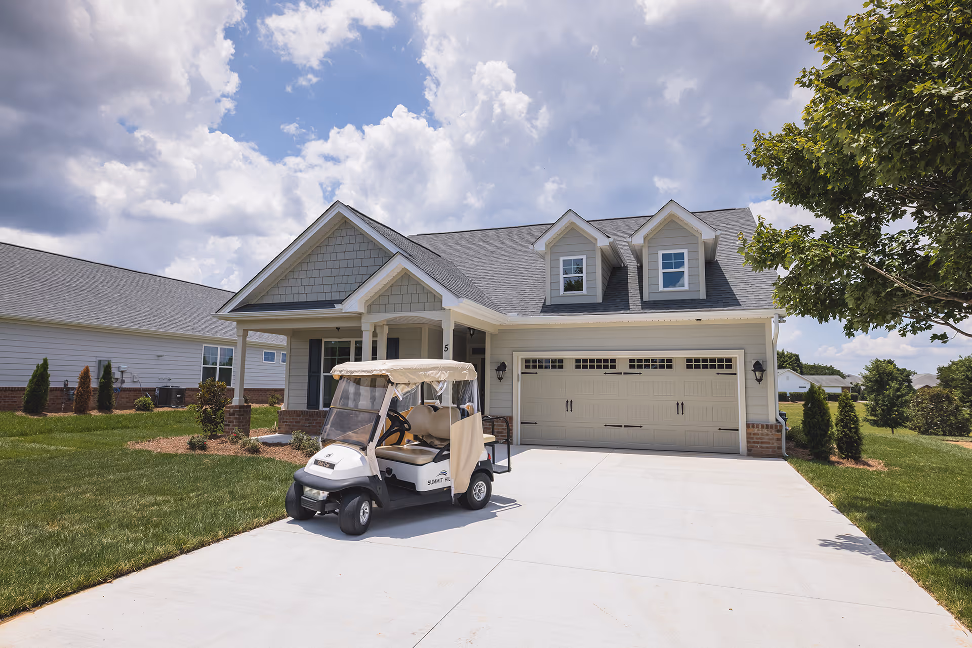 Exterior view of a single-story residential building with a two-car garage and a covered front porch. A golf cart is parked on the driveway in front of the garage. The sky is partly cloudy, and there is a tree on the right side of the driveway with green grass and landscaping around the house.