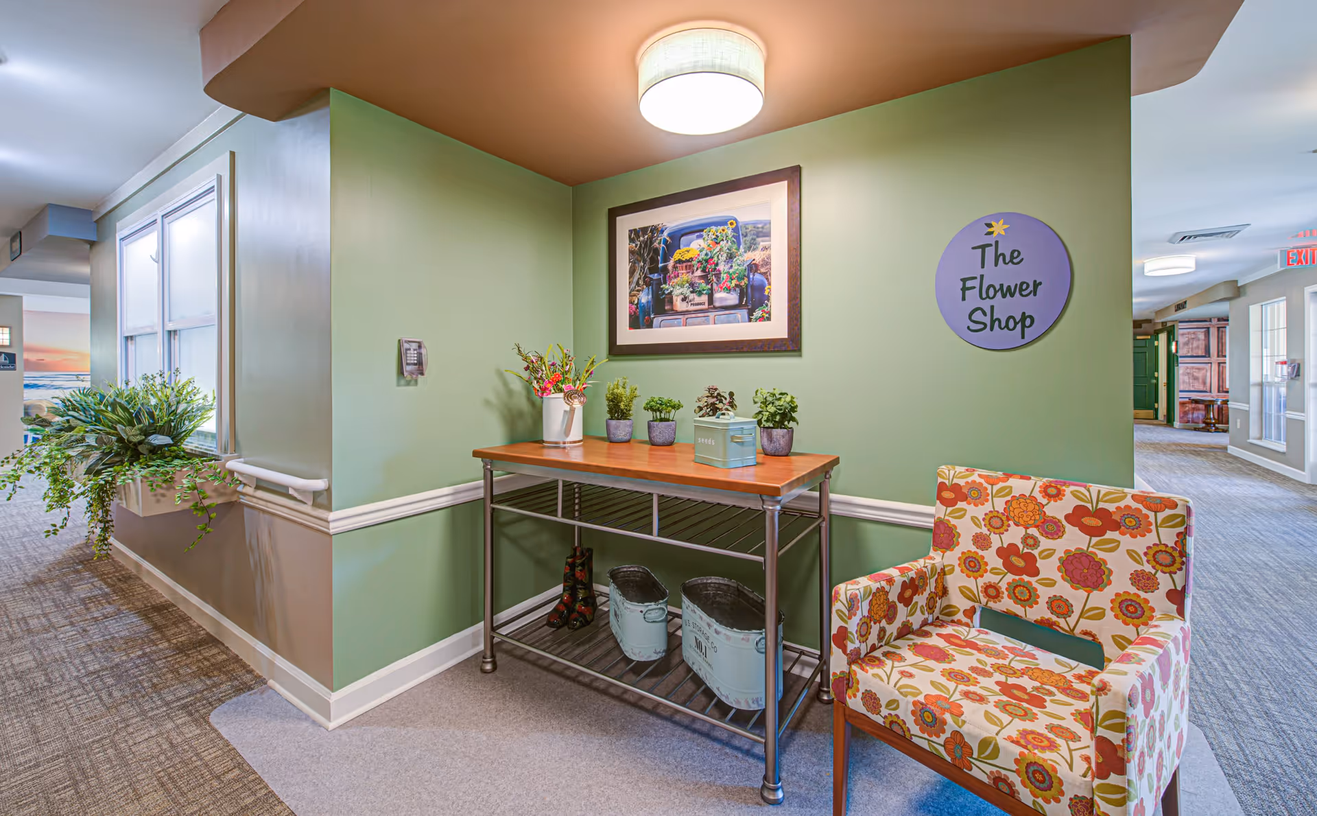 A cozy corner in a senior living community hallway featuring a green wall with a framed picture of flowers, a wooden table with small potted plants and decorative items, a floral patterned armchair, and a round sign that reads 'The Flower Shop'. The hallway has carpeted floors and light green and beige walls with white trim.