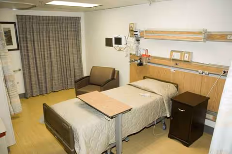 A hospital patient room with bed, chair, overbed table, and wall-mounted medical equipment.