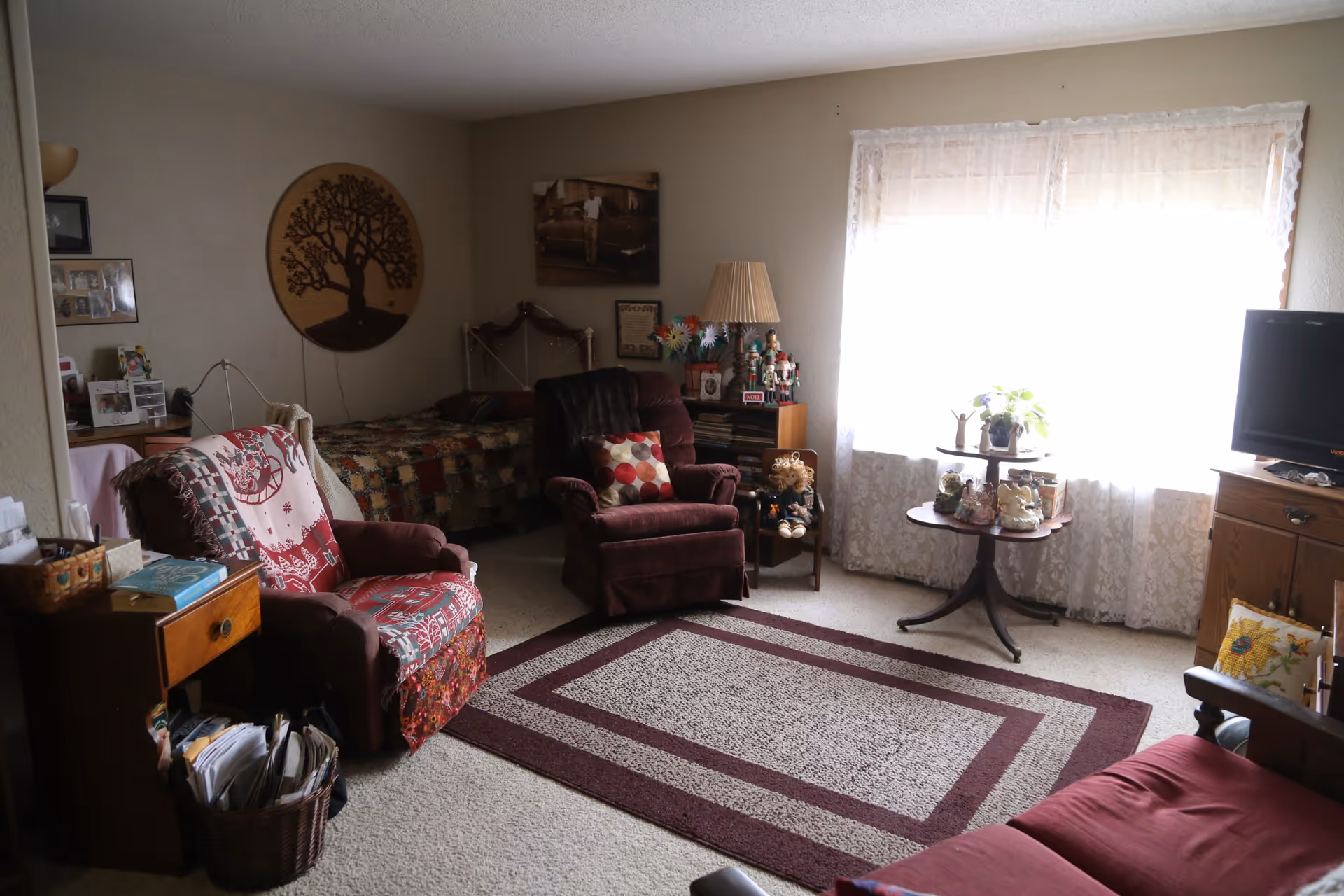 Comfortable living area with upholstered chairs, a patterned rug, side tables and a TV, with a bed and wall art visible in the background.